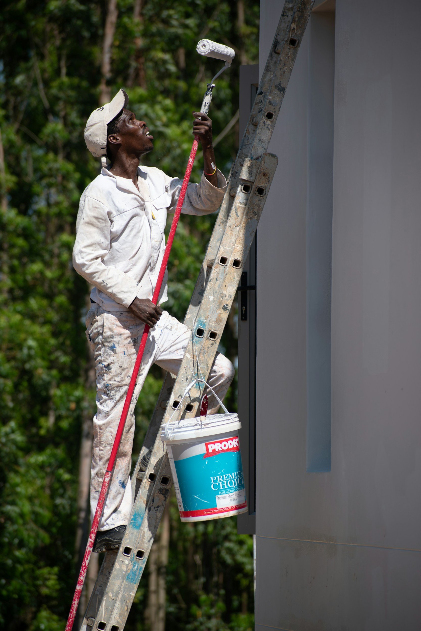 A man standing on a ladder painting a wall white with a paint roller, holding a bucket of ProDec brand paint, outdoors with a background of trees.