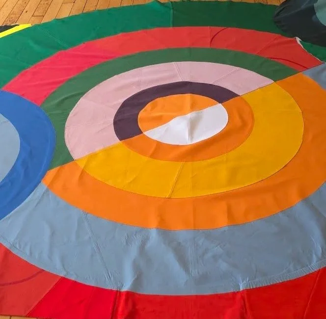 Colourful concentric circles made from reused fabrics. A close-up of a room-sized dress covered in bright, abstracted, harmonious, and clashing tones. Part of the dress that lies flat on the ground.