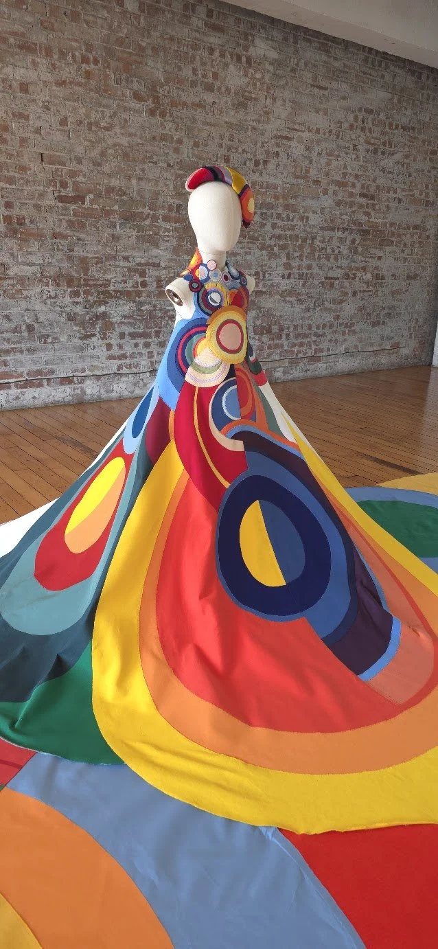 Colourful dress sculpture on a child mannequin, featuring abstract patterns with bright, swirling shapes in red, yellow, blue, and green, displayed in a gallery and taking up the majority of the room