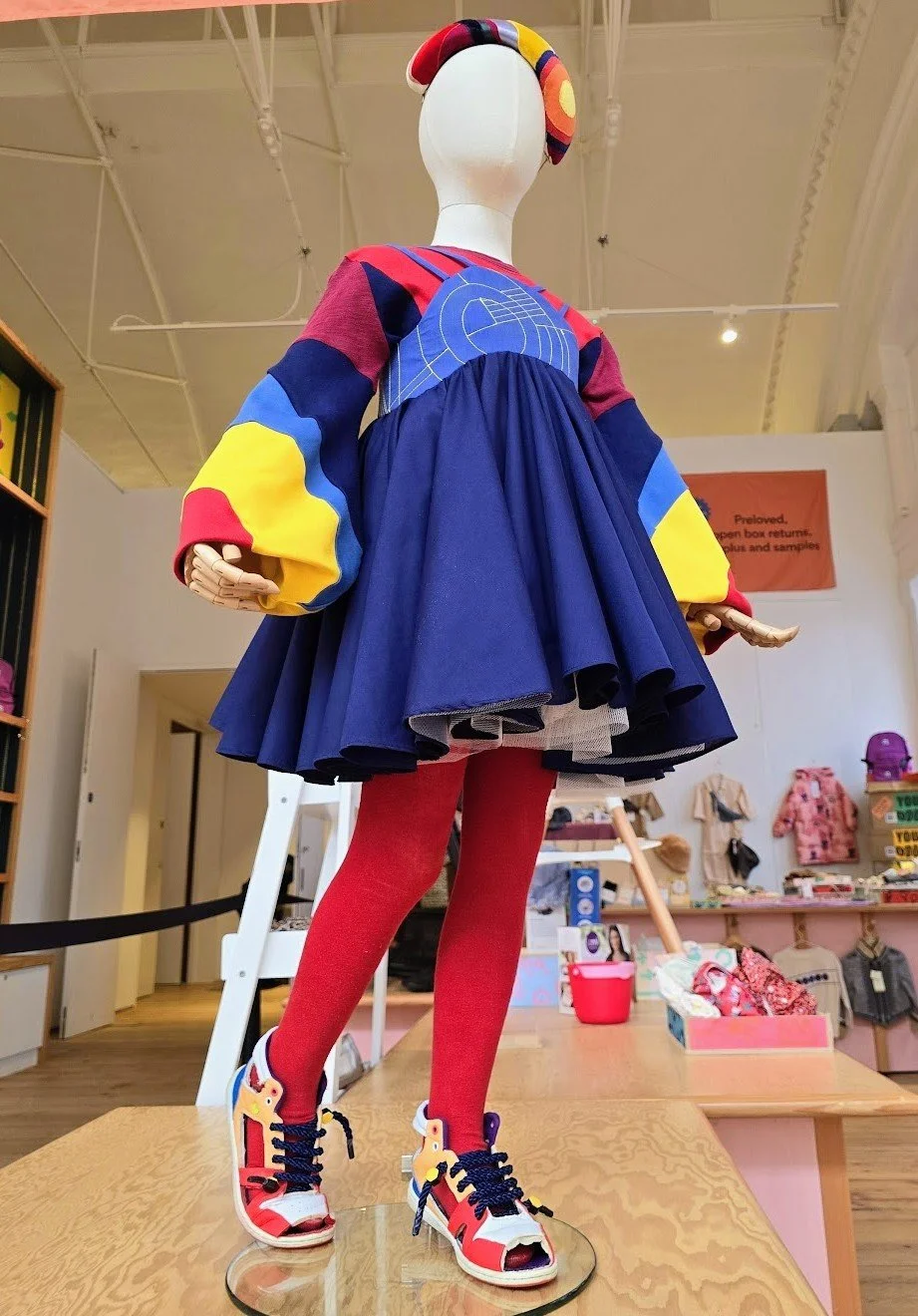 Colourful mannequin dressed in a reused textile primary colour outfit with an abstract embroidered top, a blue gathered dress with bright red tights, and bold primary coloured cutaway sneakers featured as the display design in the Young V&A Museum