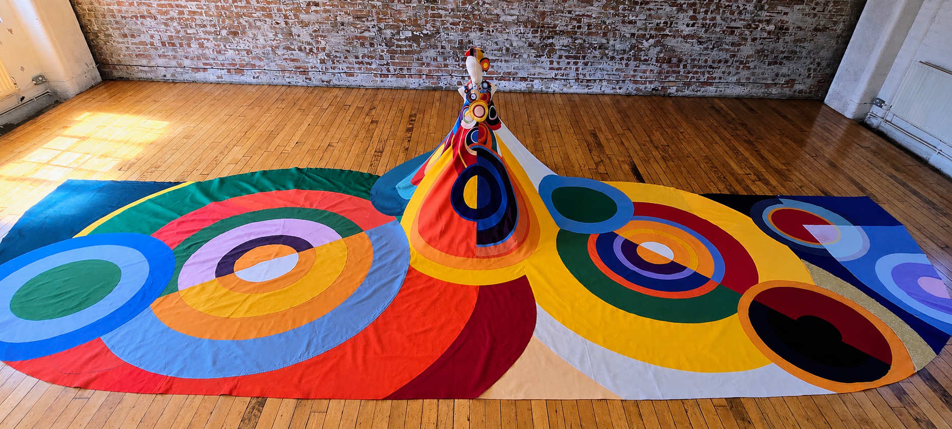 Large-scale abstract art piece by Generation Upcycle. A colourful, room-sized dress made of reuse fabric with bold concentric circles on a central mannequin, displayed on a wooden floor in a large gallery.