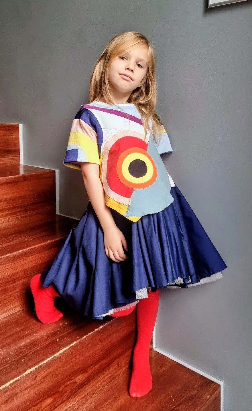 A young girl with long blonde hair wearing a colorful, abstract-patterned T-shirt, a dark blue pleated skirt, and bright red socks, standing on wooden stairs against a gray wall.