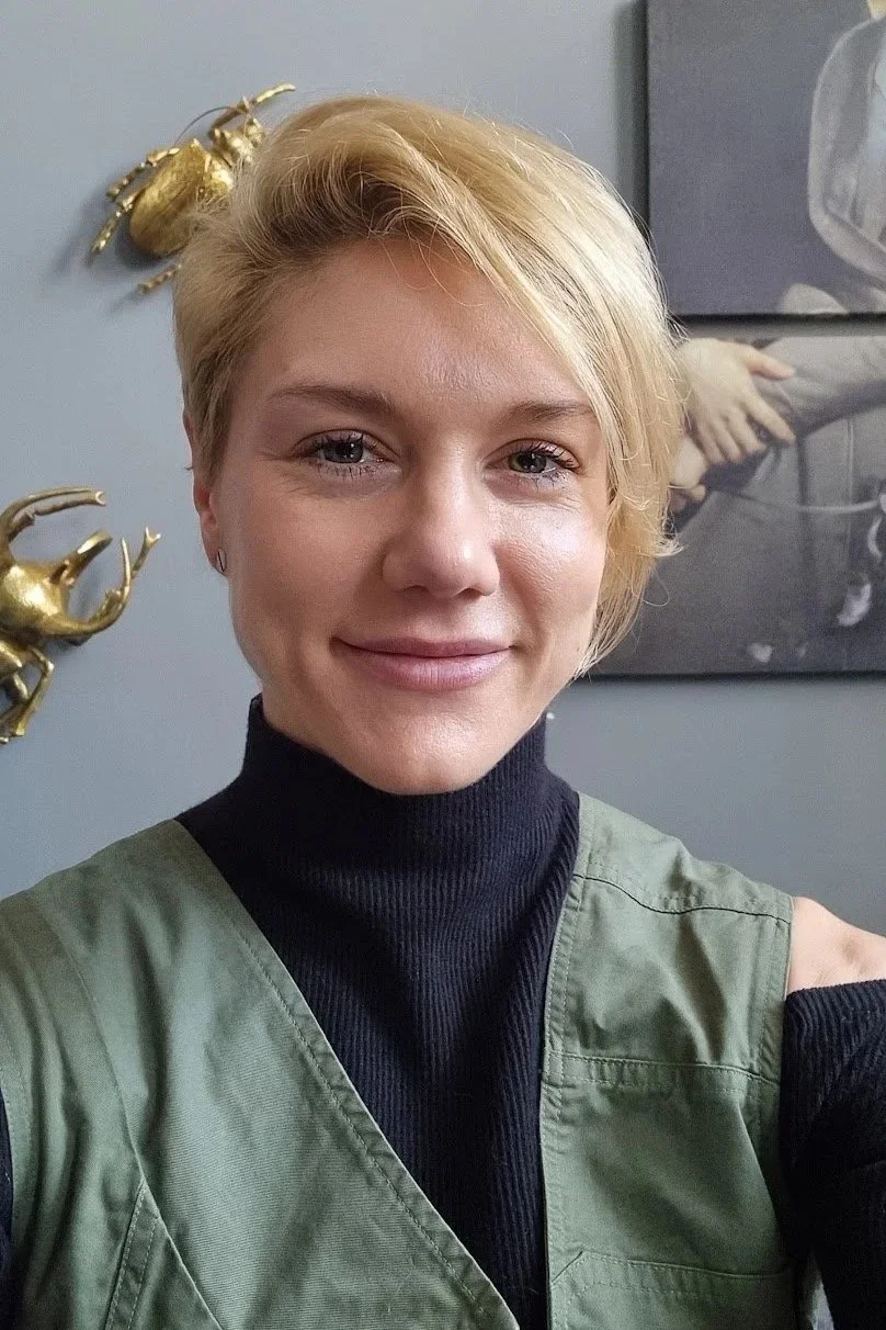 Brett Mettler, a female artist with short blonde hair, smiling at the camera, wearing a black turtleneck and upcycled green vest, with decorative beetle wall art and a black-and-white photograph in the background.