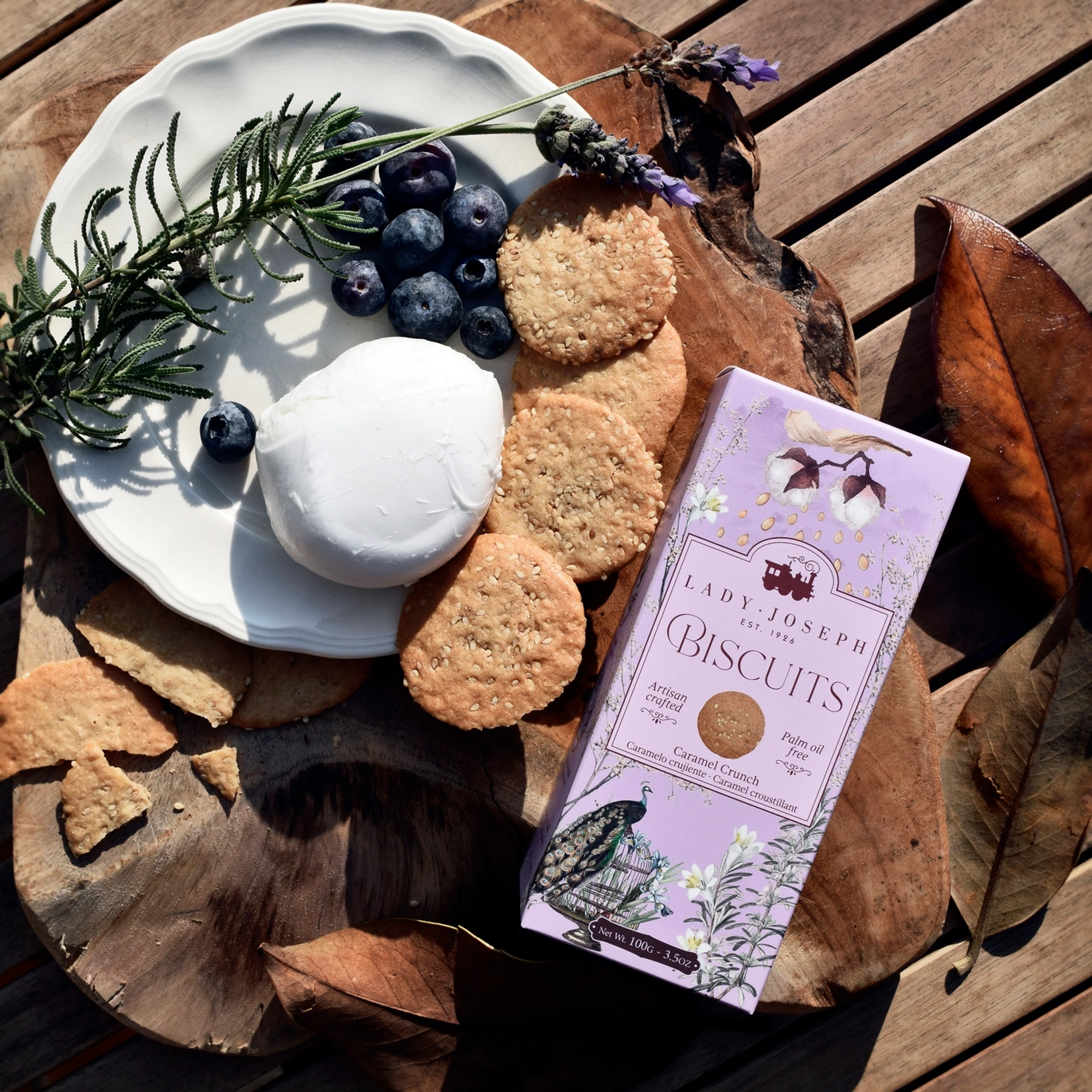 Biscoitos de Caramelo e Sésamo Lady Joseph, com embalagem lilás, apresentados numa tábua de madeira junto de uma sobremesa e frutos.