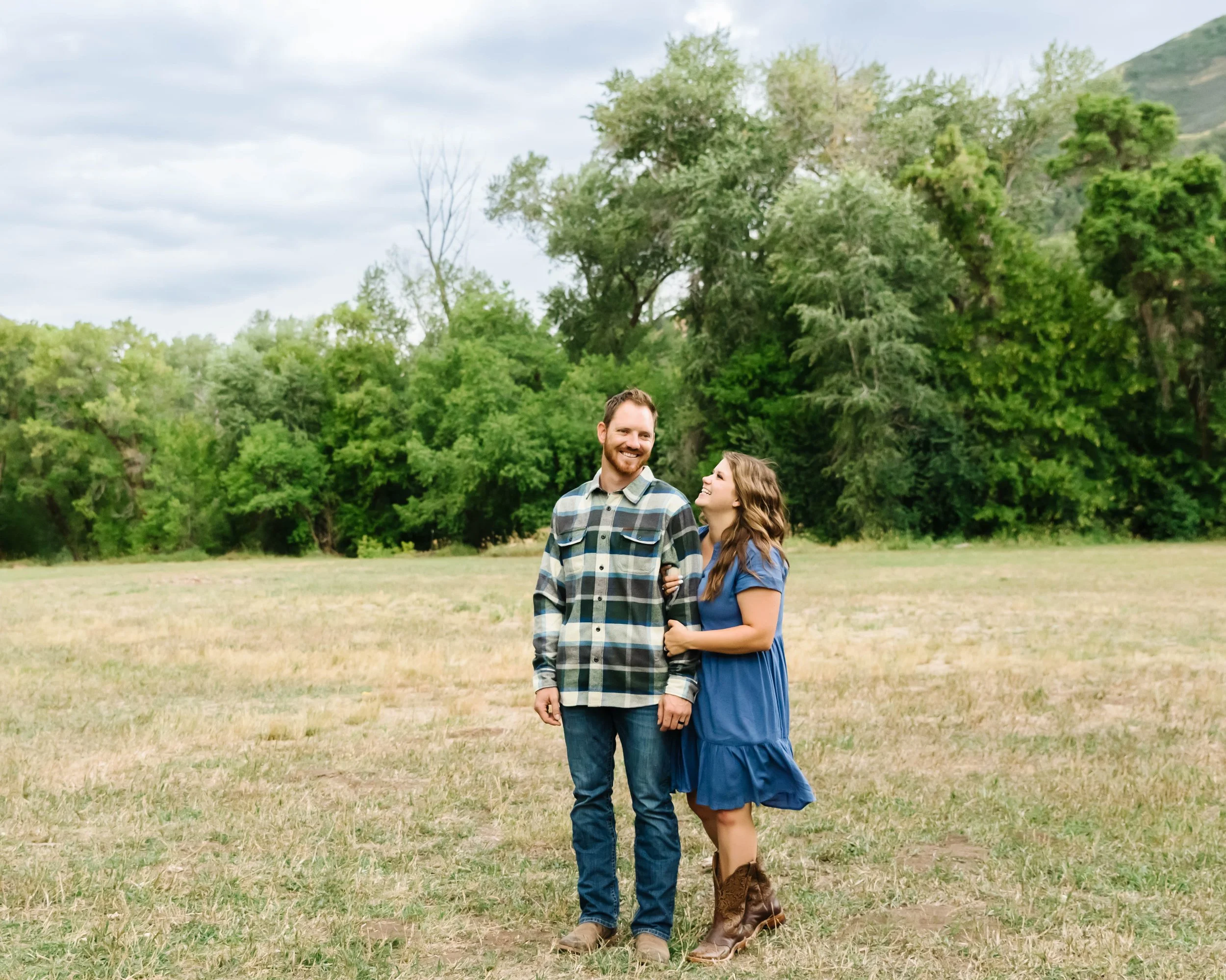 A smiling couple walking together outdoors in a grassy field with lush green trees and rolling hills in the background on a cloudy day.