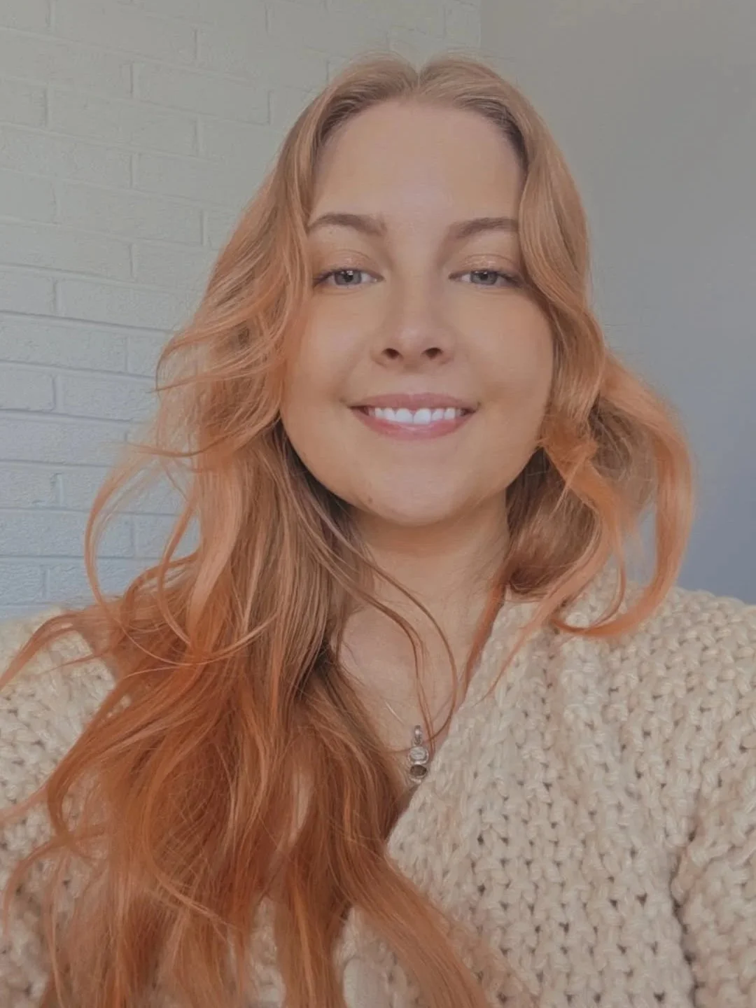 A woman with long, wavy, reddish-orange hair smiling at the camera, wearing a beige cardigan and a necklace, in front of a light-colored brick wall.