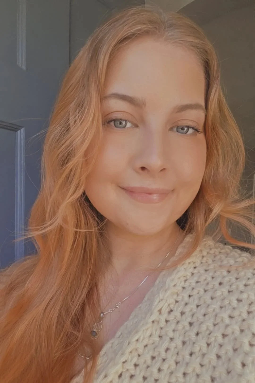 A young woman with long, wavy red hair and light blue eyes, smiling gently, wearing a beige knitted sweater and a delicate necklace, standing in front of a door.