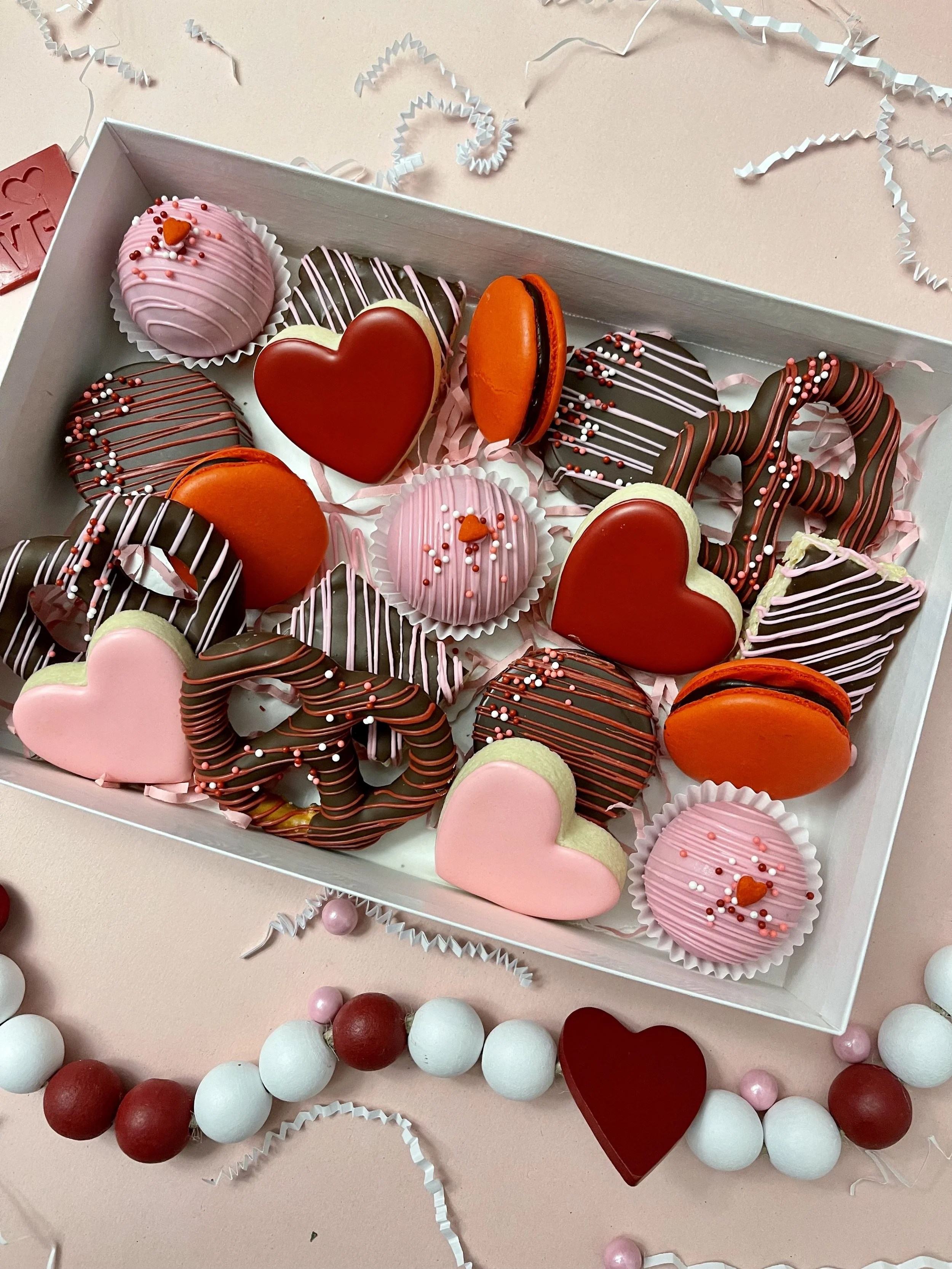 A white box of assorted decorated cookies shaped like hearts and circles, with pink, red, brown, and orange icing, placed on a pink surface with pink, white, and maroon beads and paper confetti around it.