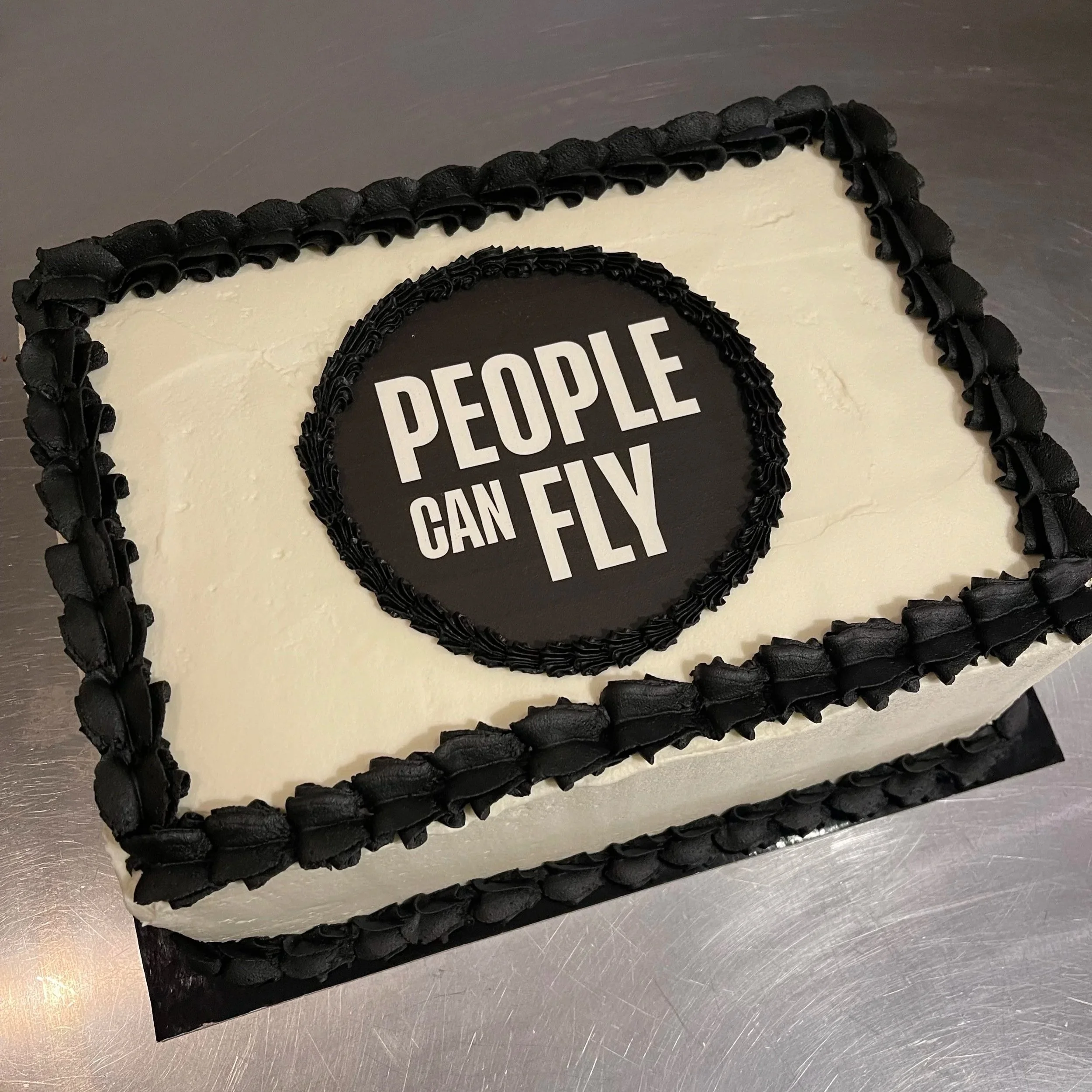 A cake decorated with black and white frosting, with the phrase 'PEOPLE CAN FLY' on the top. The cake has a black ruffled border around the top edge and perimeter.