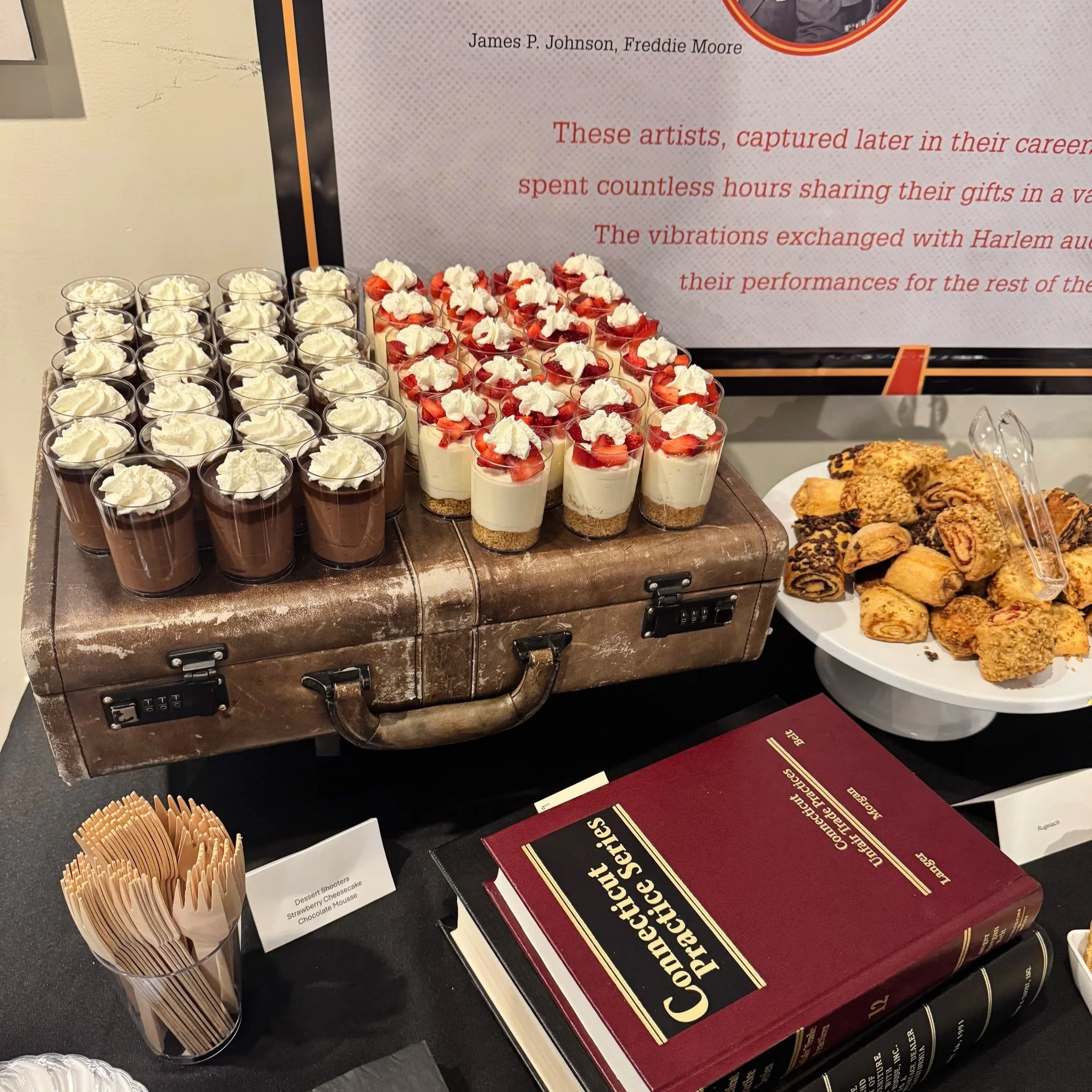 An assortment of desserts including chocolate mousse and strawberry cheesecake cups on a table next to a guide book and a container of uncooked spaghetti.