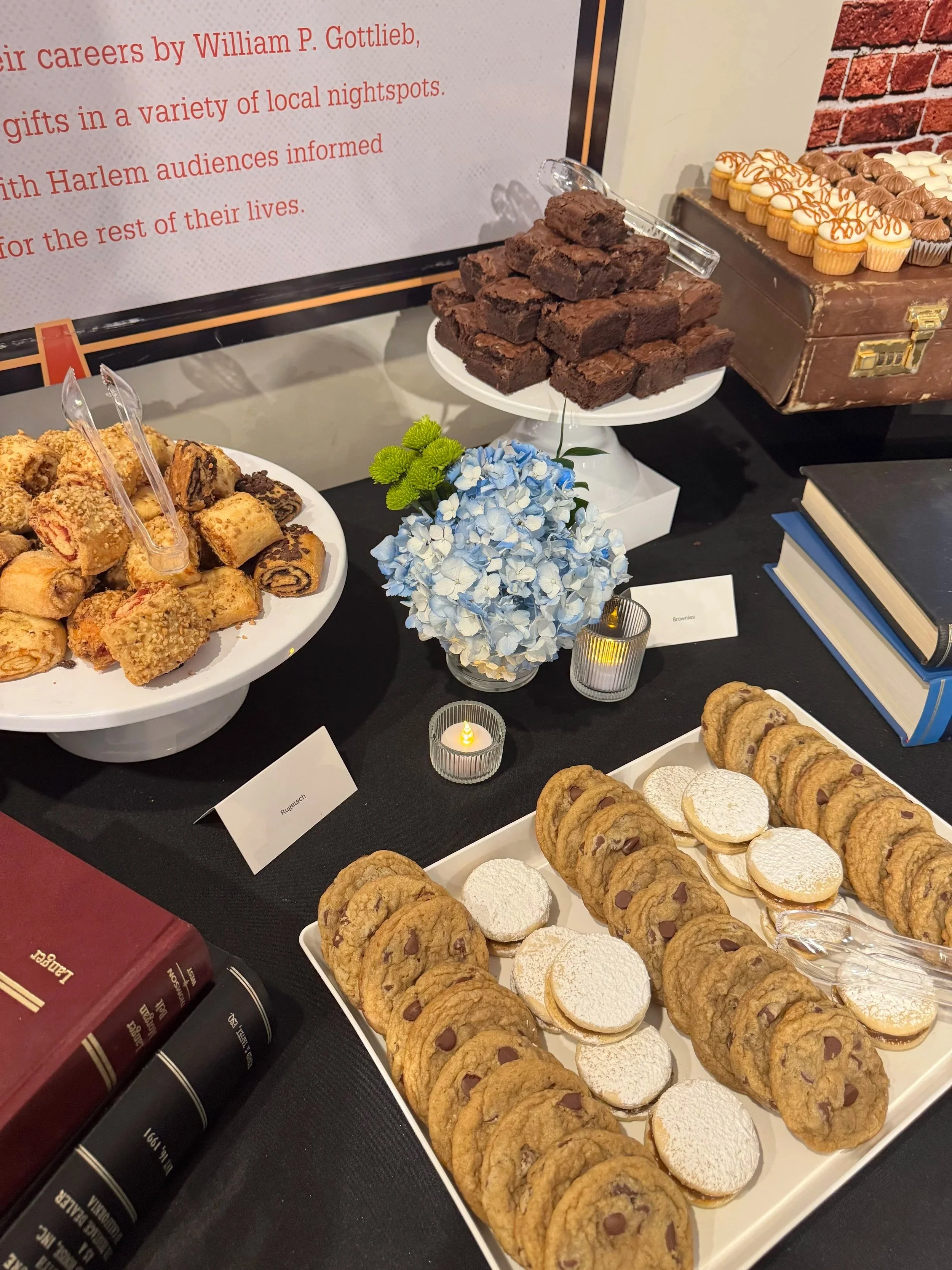 Display of various desserts including a plate of cookie sandwiches with chocolate chips and powdered sugar, a tray of layered cookies with chocolate chips, a bunch of chocolate brownies, and a tray of cupcake-like confections with white, chocolate, a