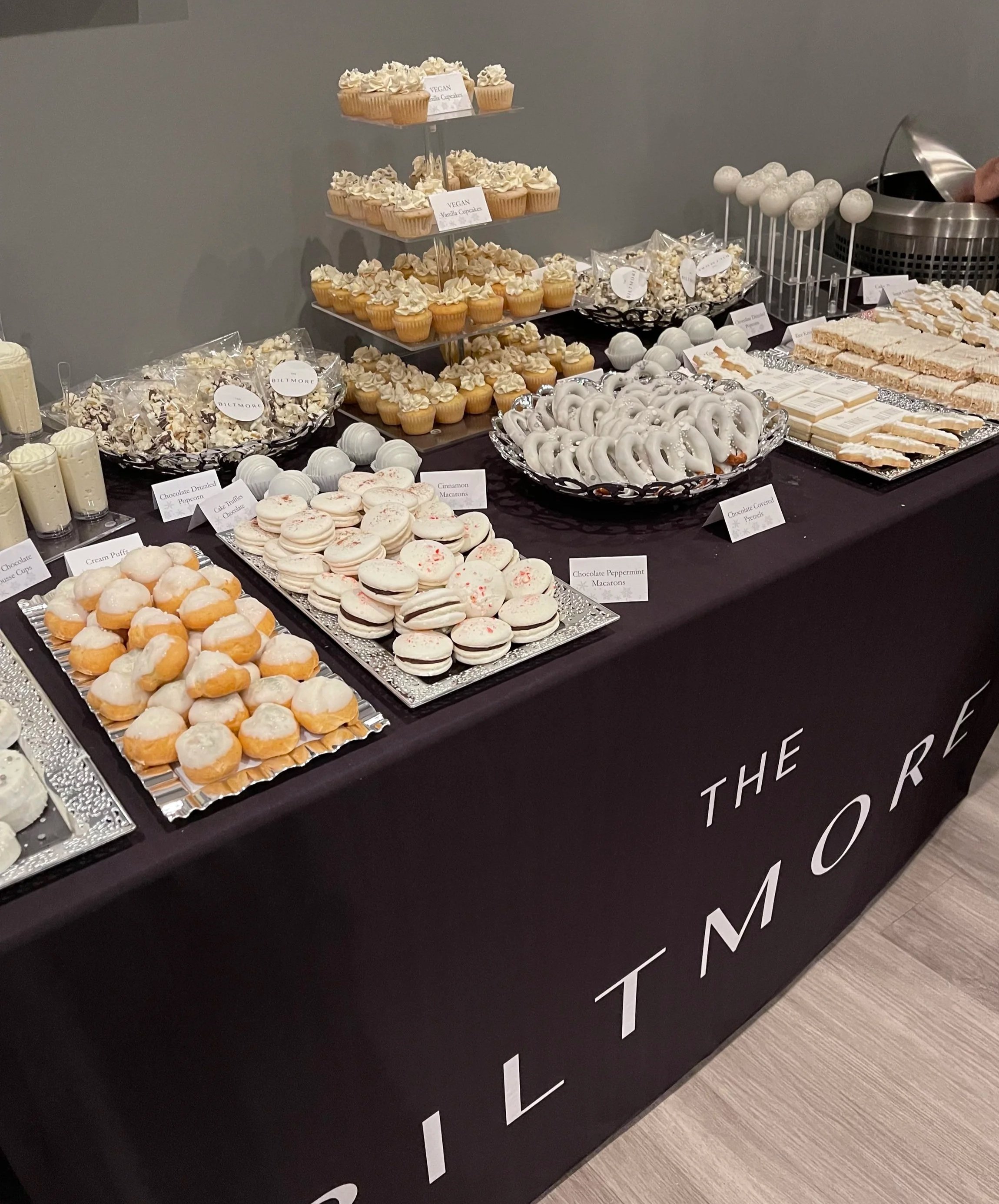 A dessert table with various sweet treats, including cupcakes, macarons, cake pops, and popcorn, all decorated in white and beige, with labels indicating flavors like vanilla, chocolate, and peppermint, set against a gray wall.