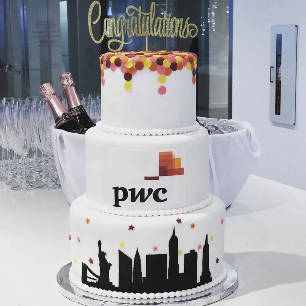 Three-tiered celebration cake with confetti and city skyline decorations, branded with PwC logo, topped with a gold "Congratulations" cake topper, surrounded by champagne bottles and glasses in a festive setting.