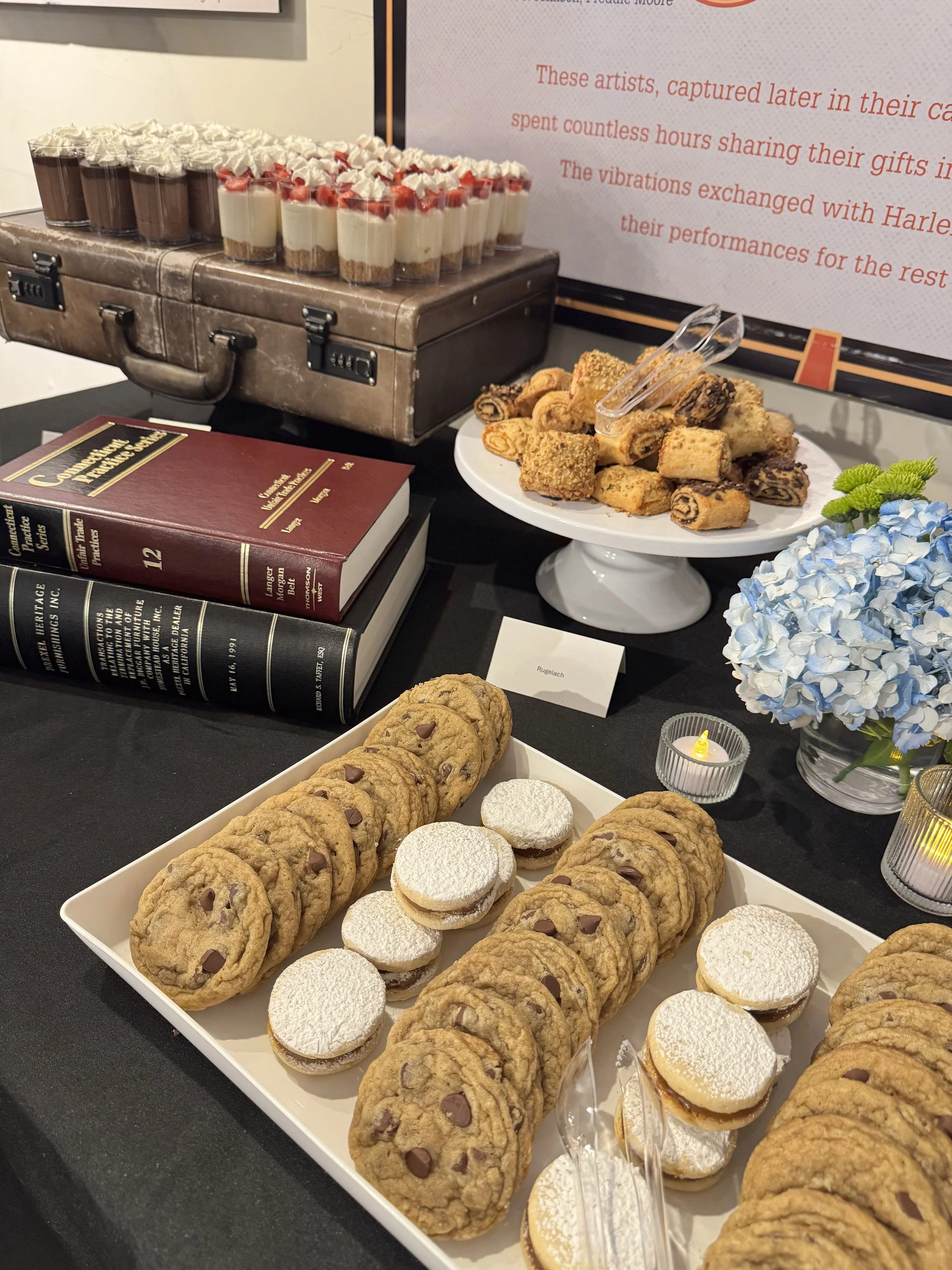 Tray of assorted cookies next to a tray of mini cheesecakes topped with powdered sugar, a plate of cake roll slices, and a plate of layered dessert cups with whipped cream and strawberries, beside a small lit candle and a blue hydrangea flower.