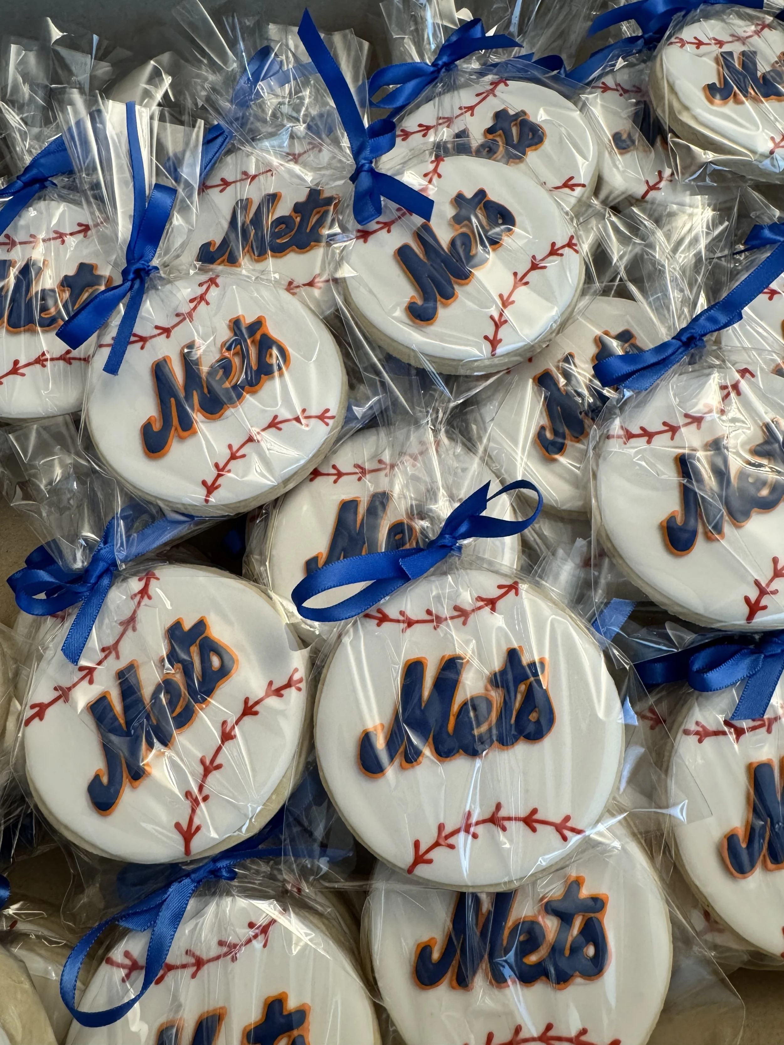 Decorated sugar cookies with blue ribbons, featuring the New York Mets logo and baseball-themed designs, wrapped in clear plastic.