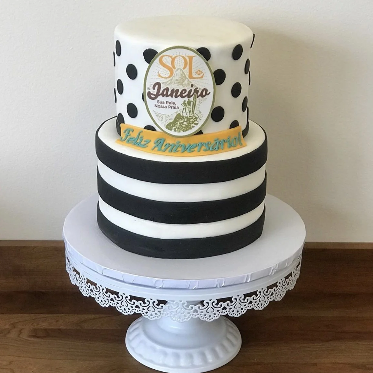 A two-tier birthday cake with black and white striped pattern on the bottom tier and a white top tier with black polka dots. The top tier has a logo with the text 'SOL Janeiro' and an illustration. A yellow ribbon with blue text 'Feliz Aniversário!' 