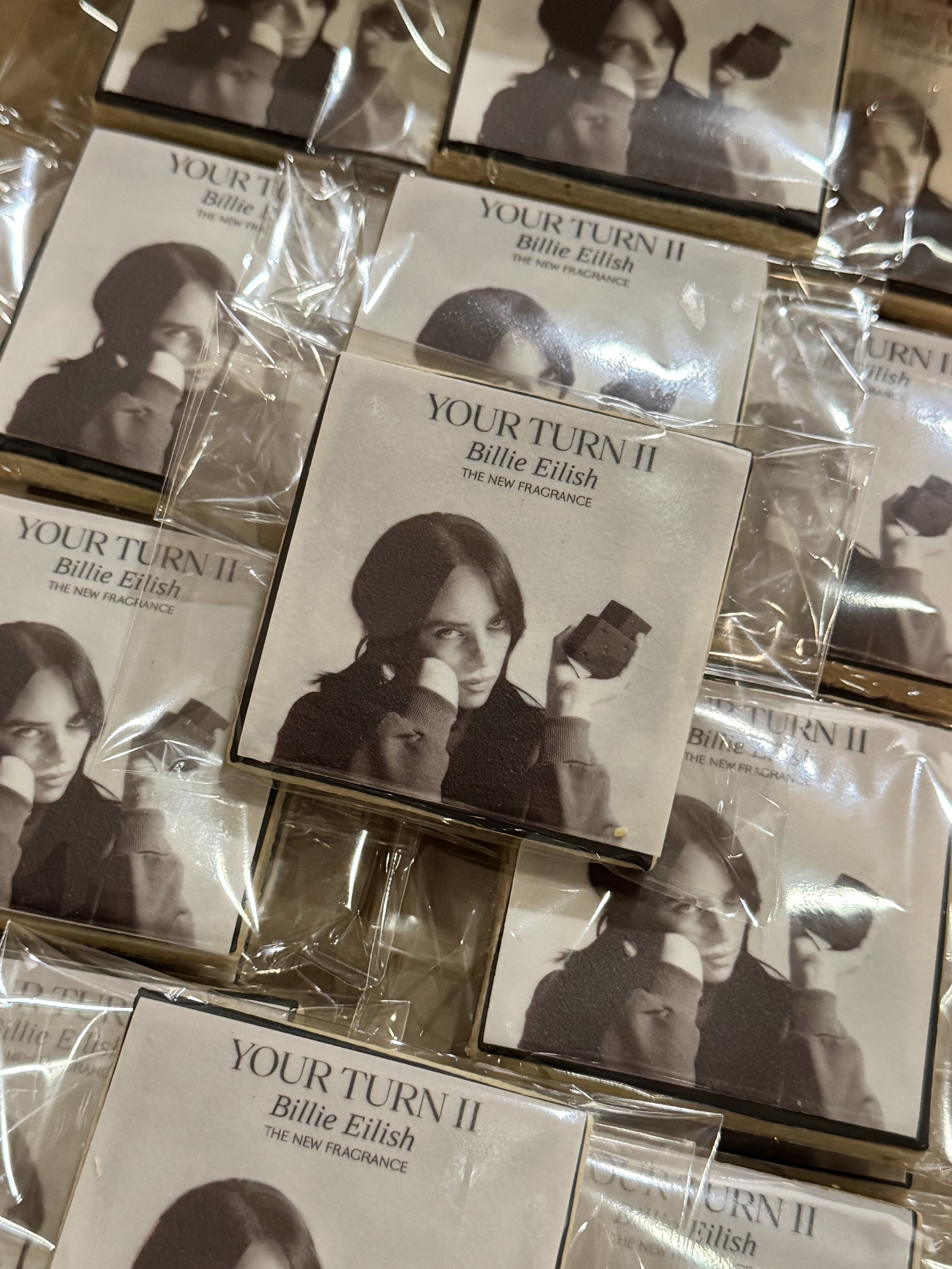 Multiple packages of Billie Eilish perfume labeled 'Your Turn II' showing a sepia-toned image of Billie Eilish holding a box of chocolates.