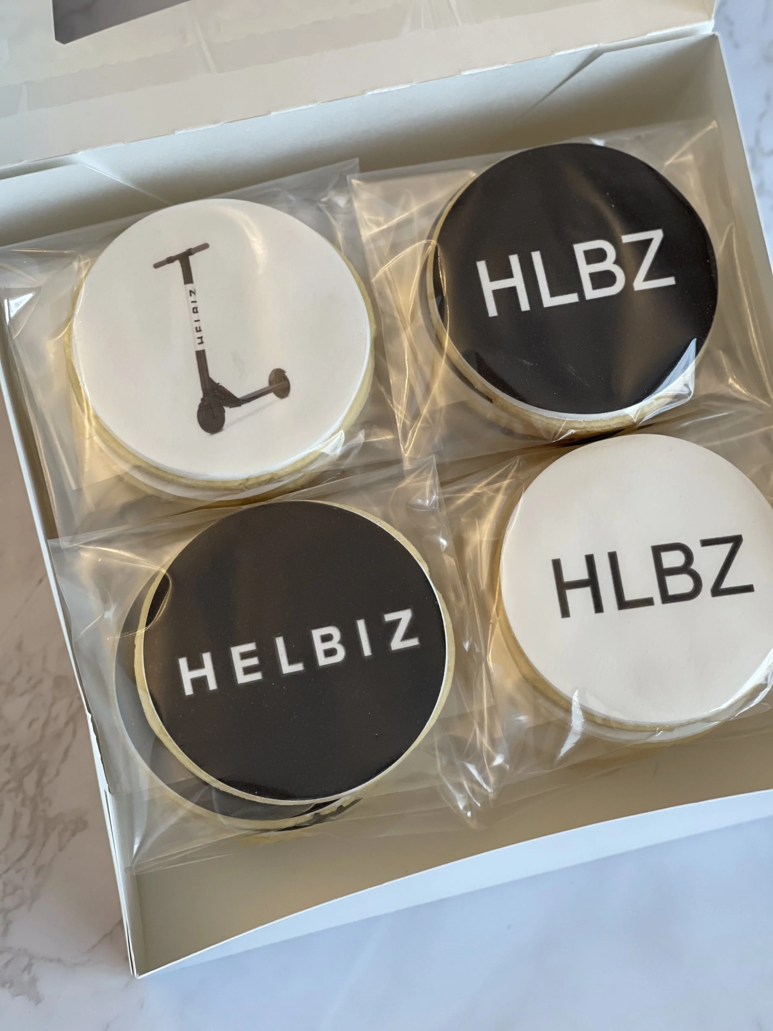 A white box with four round cookies wrapped in clear plastic, each with a different black and white design, with the word HELBIZ printed on two of the cookies.