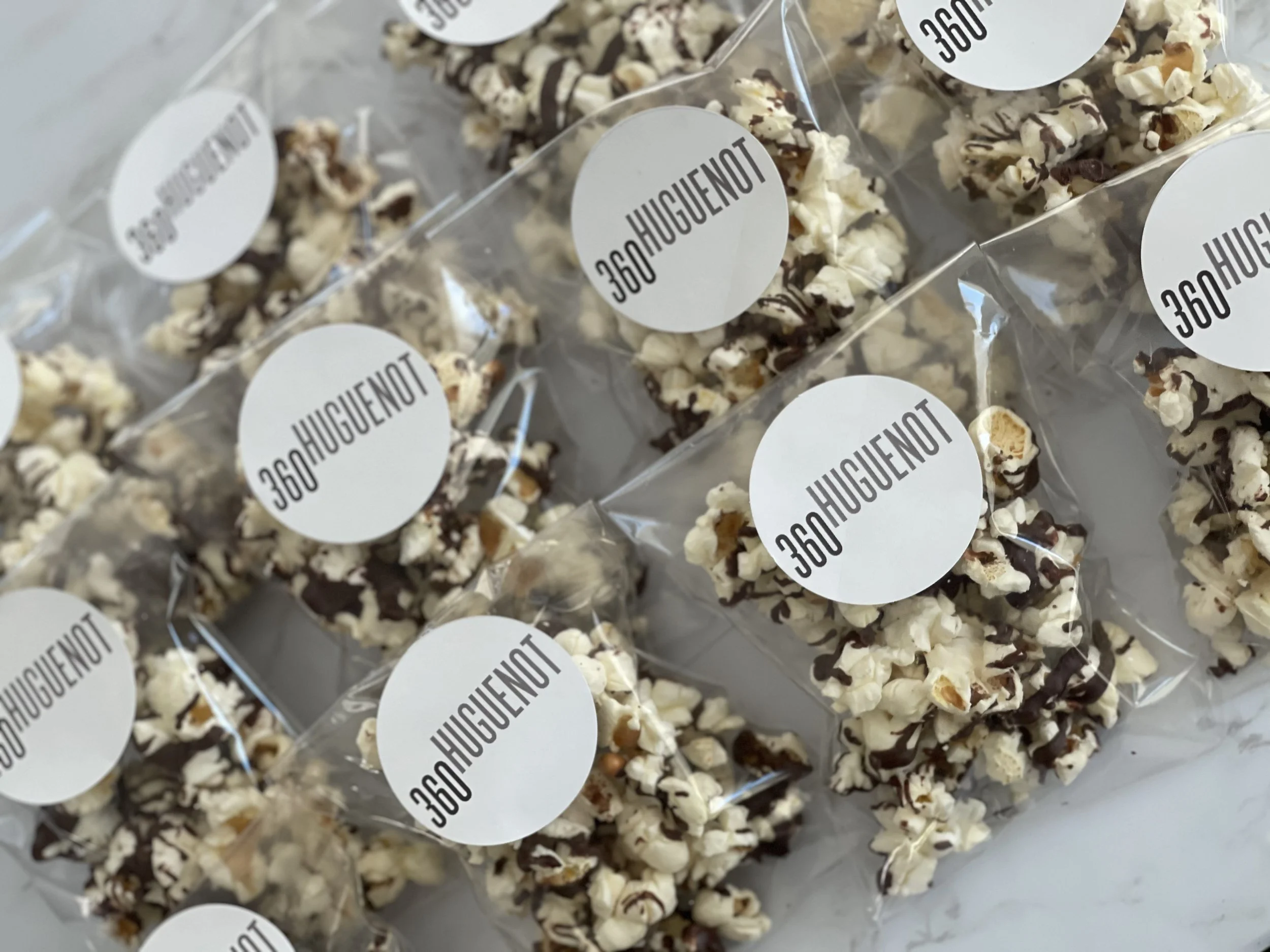 Several clear plastic bags filled with popcorn coated in chocolate and sprinkled with nuts, each with a round white label that reads '360HUGENUT'.