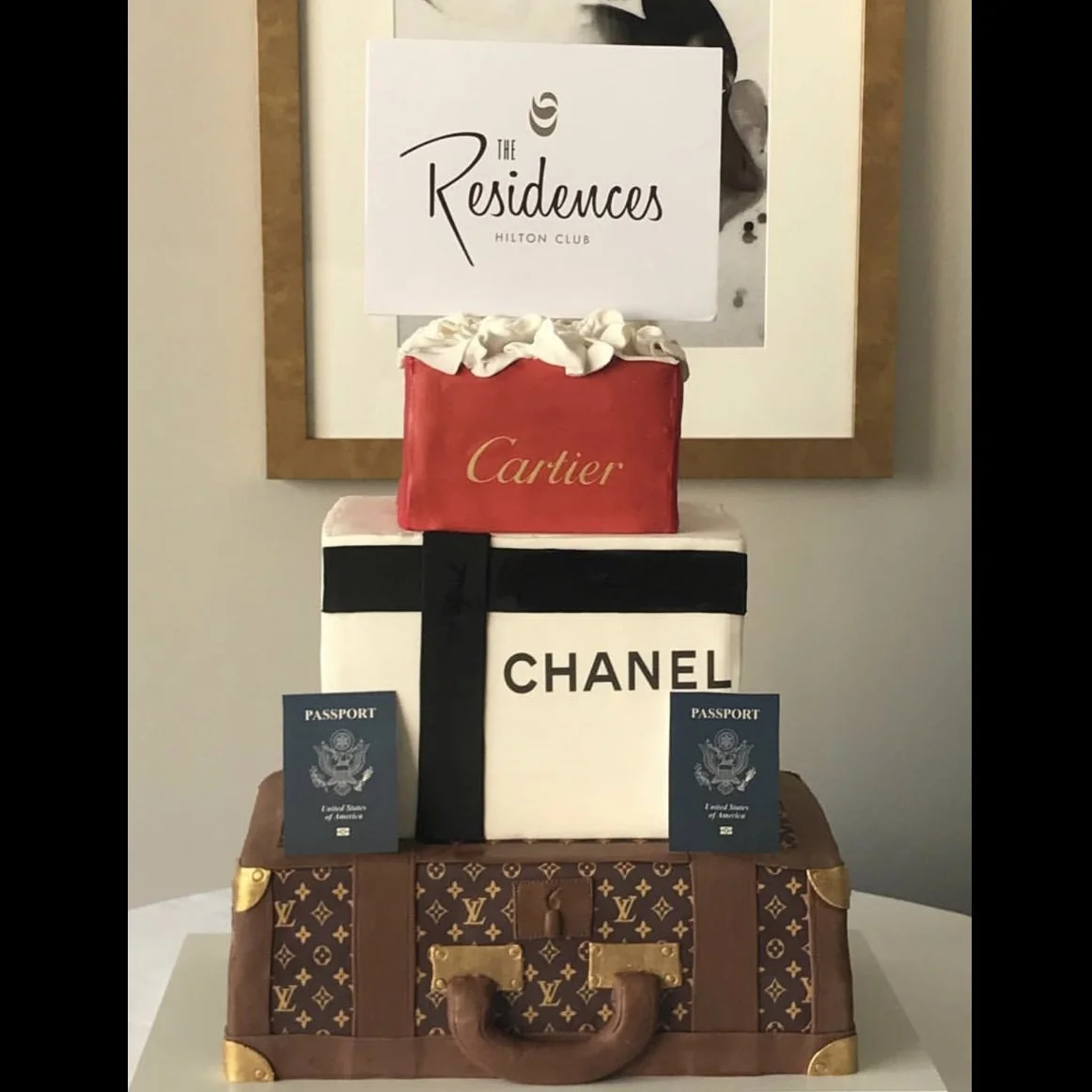 A layered display of luxury brand themed boxes, with a Louis Vuitton suitcase at the bottom, a Chanel box in the middle, and a Cartier pouch and a sign for The Residences Hilton Club at the top.