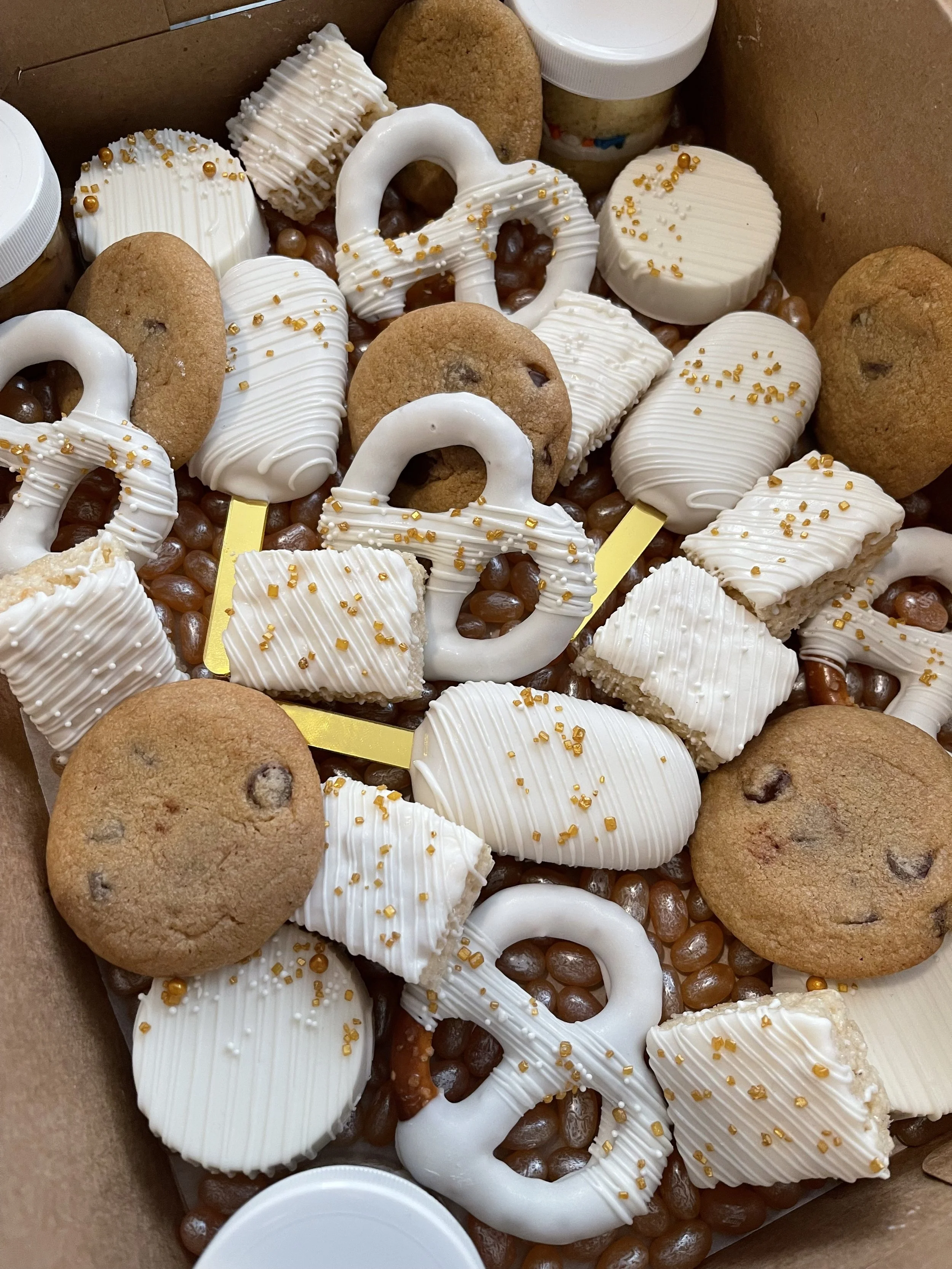 A box of assorted cookies and white icing-covered treats with gold sprinkles, surrounded by small chocolates and cookie containers.