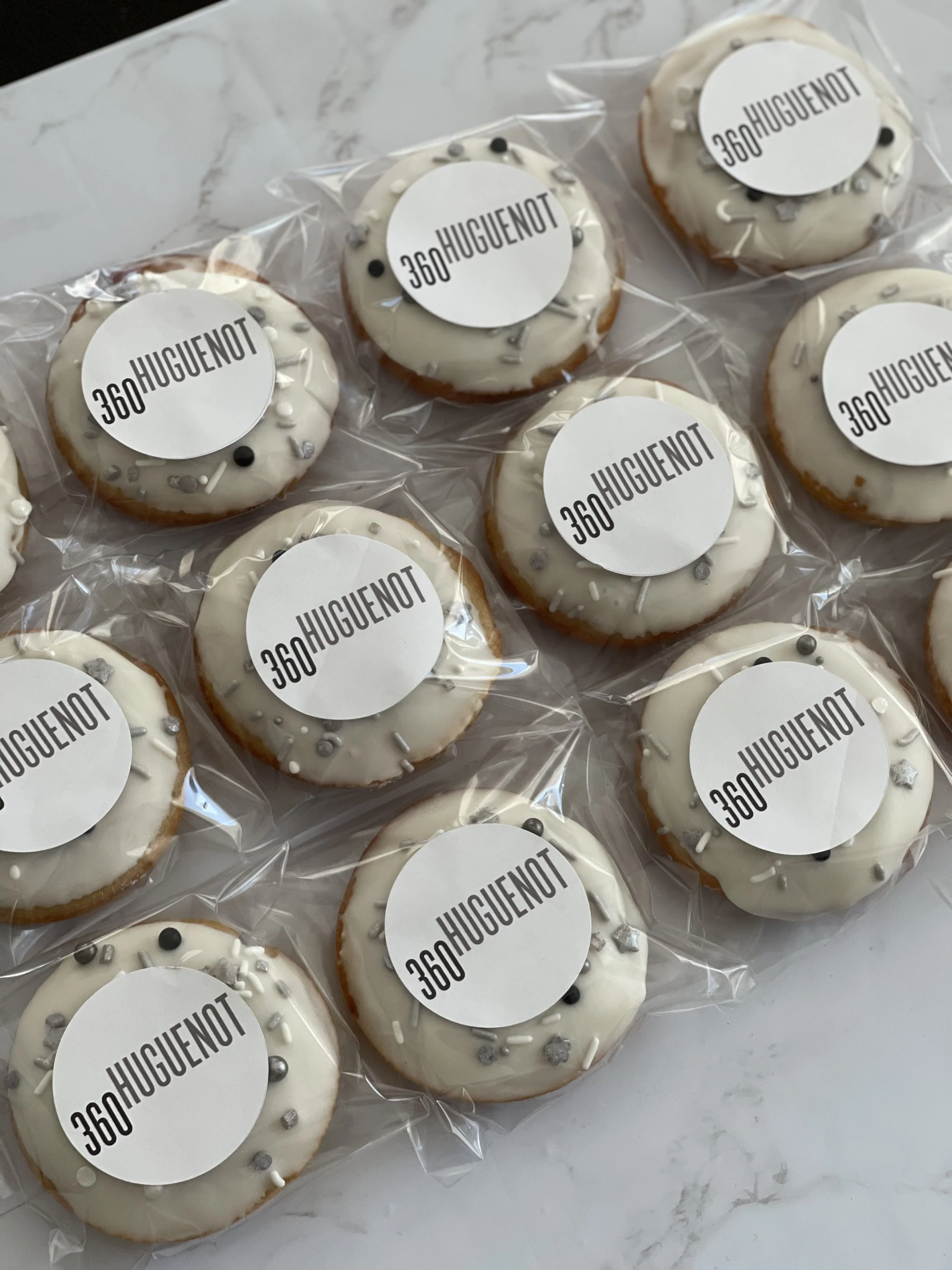 Several decorated donuts with white icing and various silver, white, and black sprinkles, each labeled with a sticker that says '360HUGUENOT' in black text.