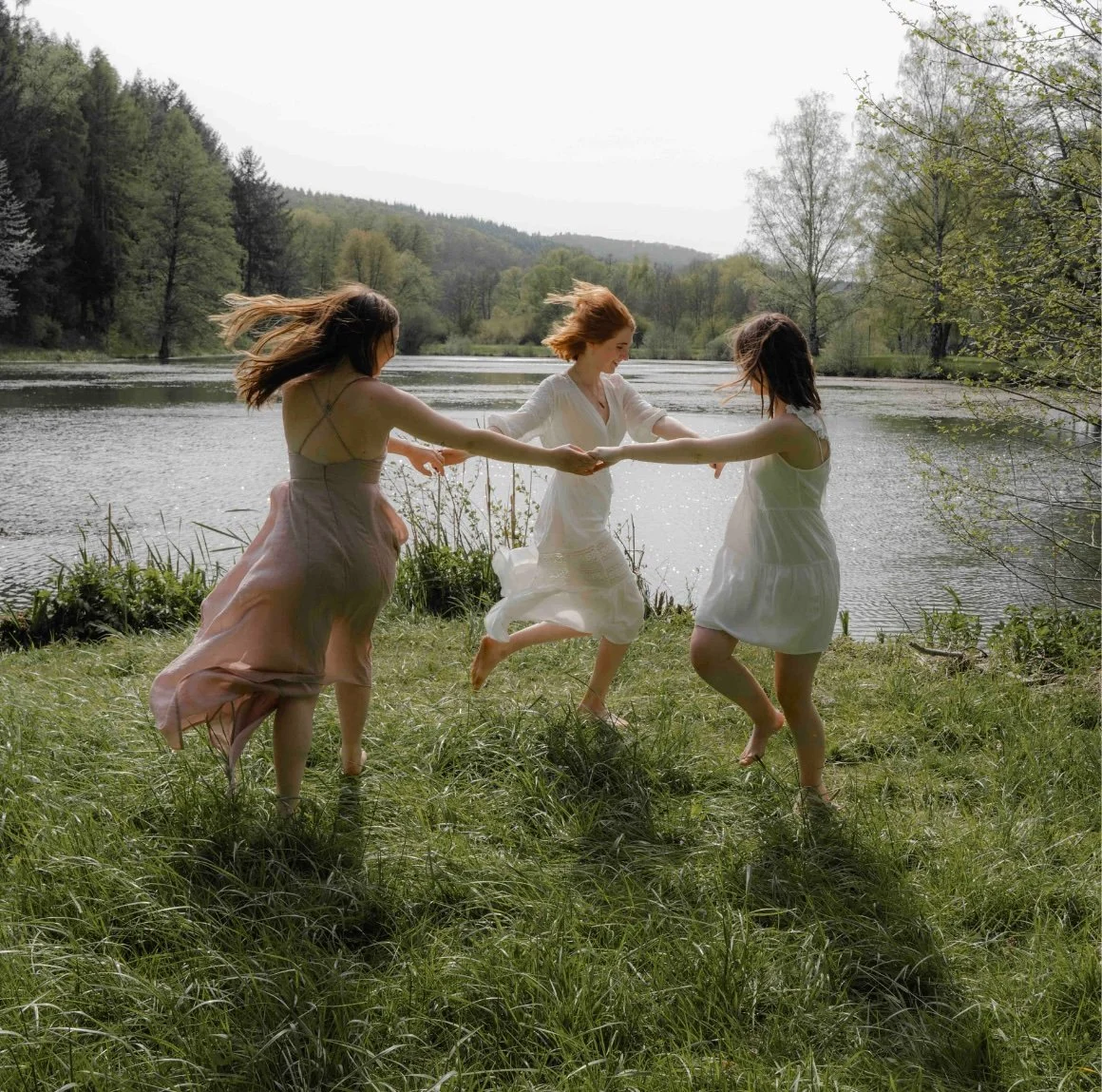 Three women in light dresses holding hands and spinning by a lakeside, surrounded by trees and grass, on a partly cloudy day.