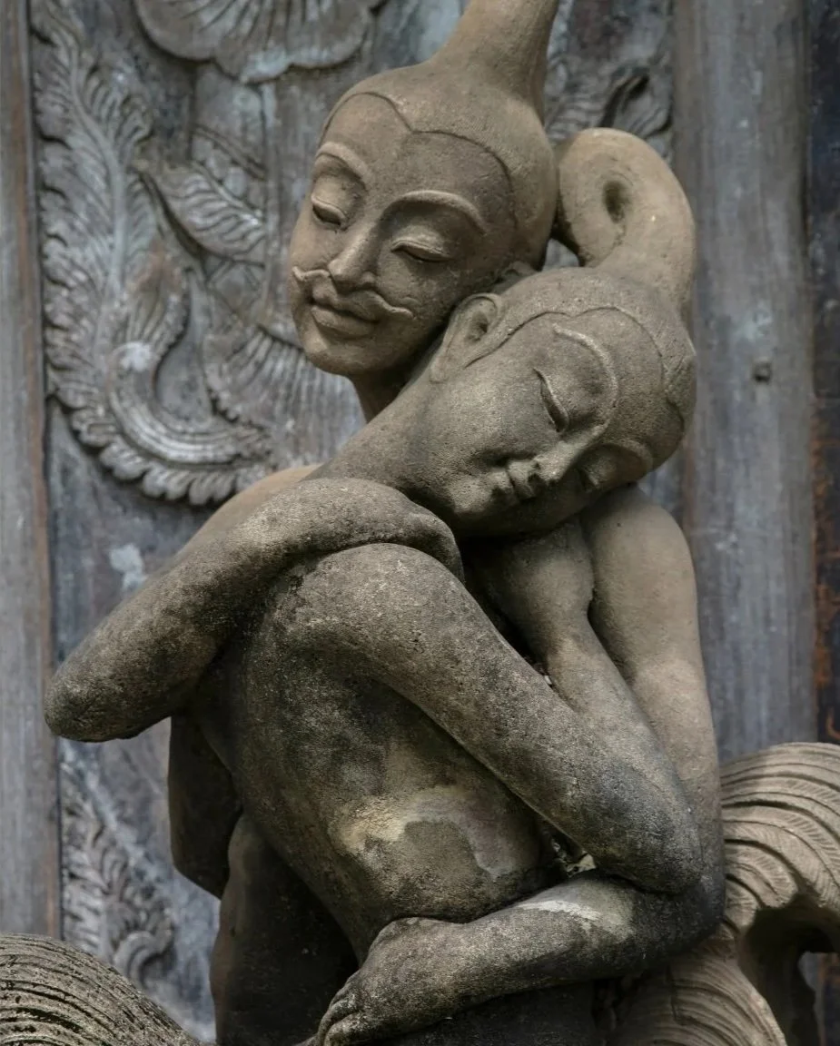 Close-up of a stone sculpture depicting two abstract human figures embracing each other, with one figure resting its head on the other's shoulder, set against a carved wooden background.
