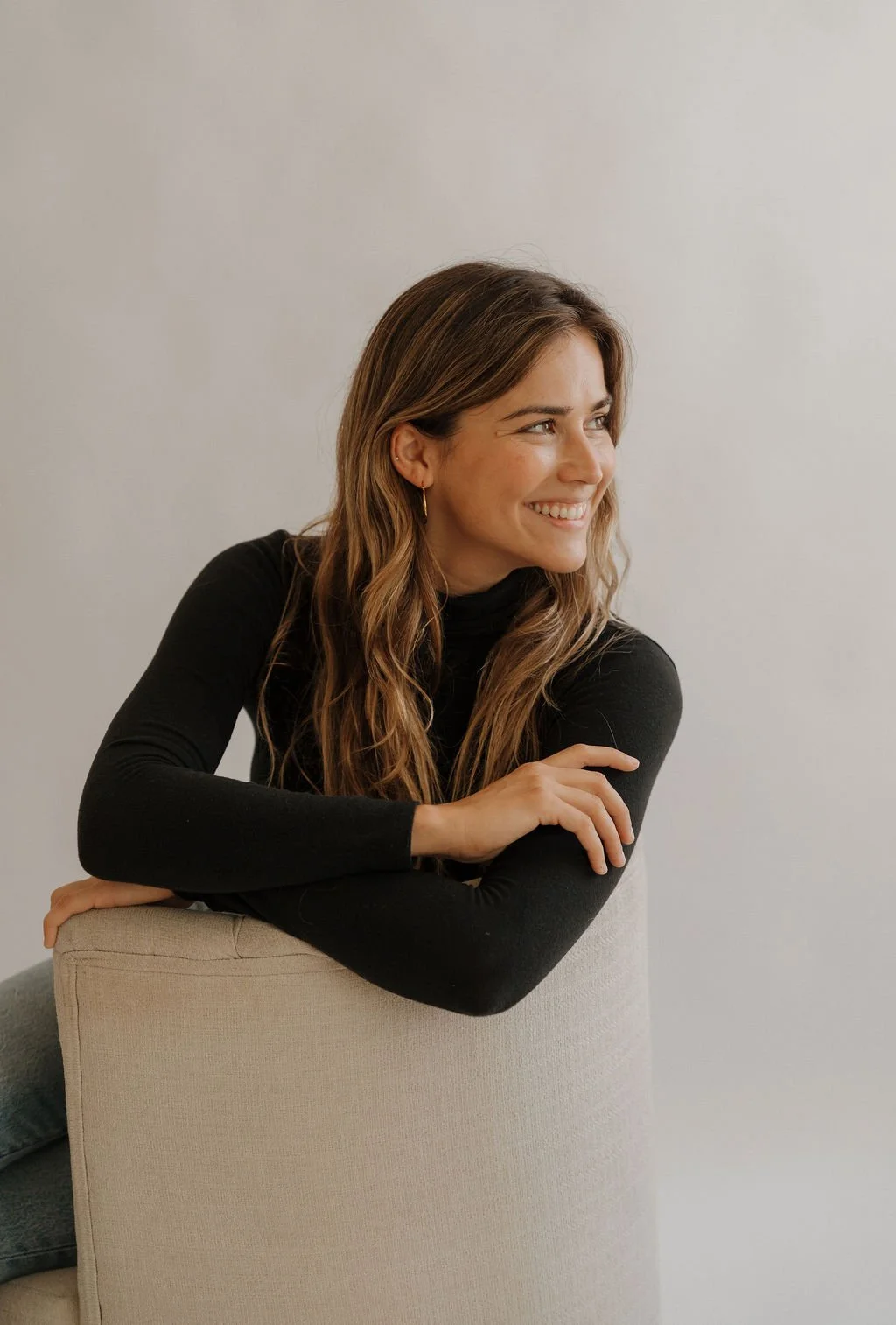 A young woman with long, wavy brown hair, wearing a black long-sleeve top, smiling and looking to her right, sitting on a beige chair against a plain wall.