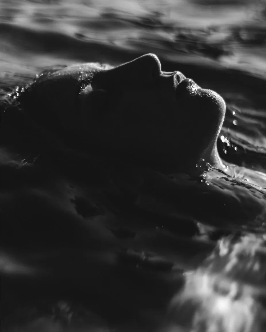 Black and white photo of a person's face partially submerged in water, viewed from the side with closed eyes.