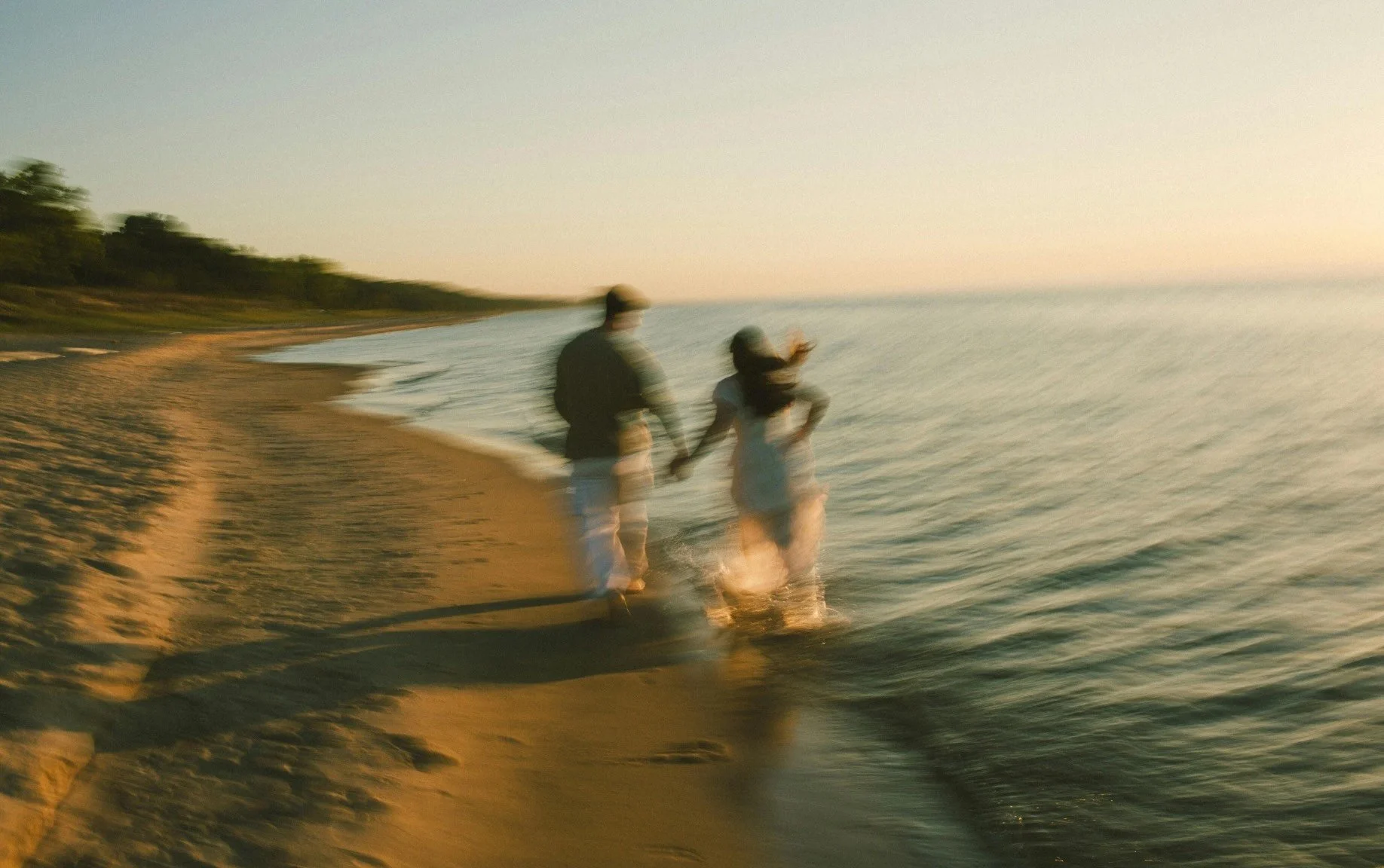 A blurry photo of a couple walking hand-in-hand along the beach at sunset, with waves and trees in the background.