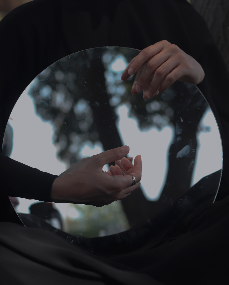 Person holding a handheld mirror outdoors, reflecting their hands and a blurred background of trees.