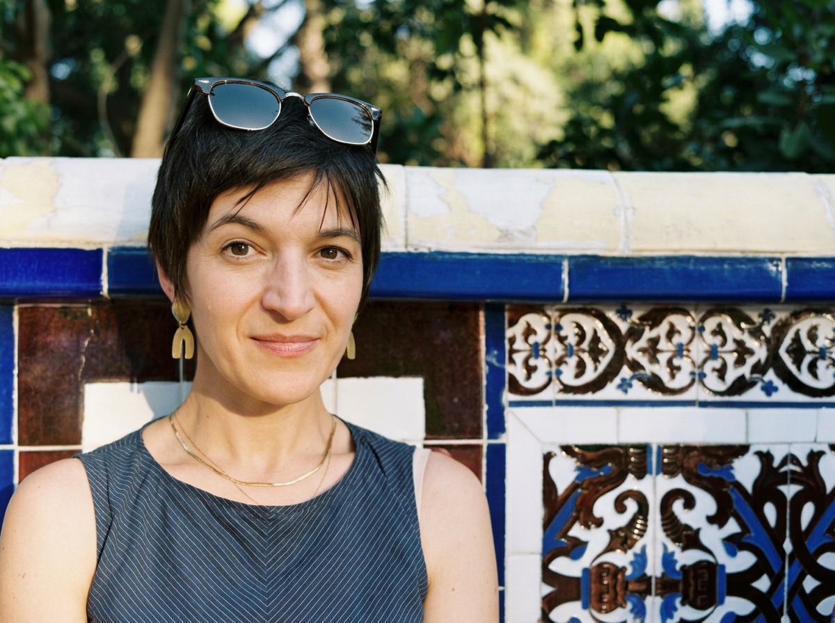 Bones Content Co. founder Cody Bay is pictured with short dark hair, wearing sunglasses on her head, gold earrings, and a sleeveless striped top, stands in front of a decorative tile wall, outdoors with blurred green trees in the background.