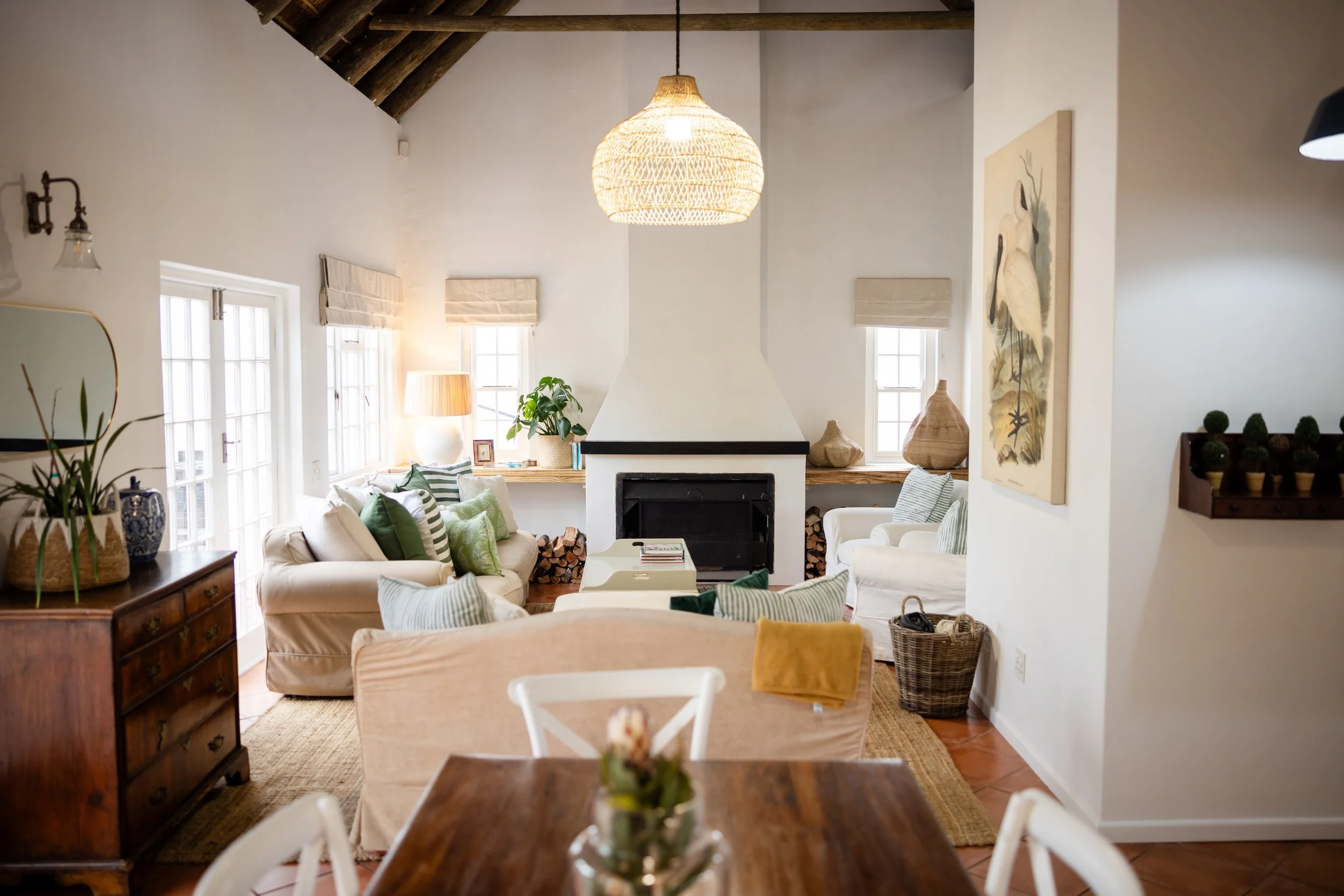 Living room with a white wall, wooden ceiling beams, wicker pendant light, white sofas with green and striped pillows, large windows with Roman shades, fireplace, large painting of a bird, potted plant, wooden furniture, and woven baskets.