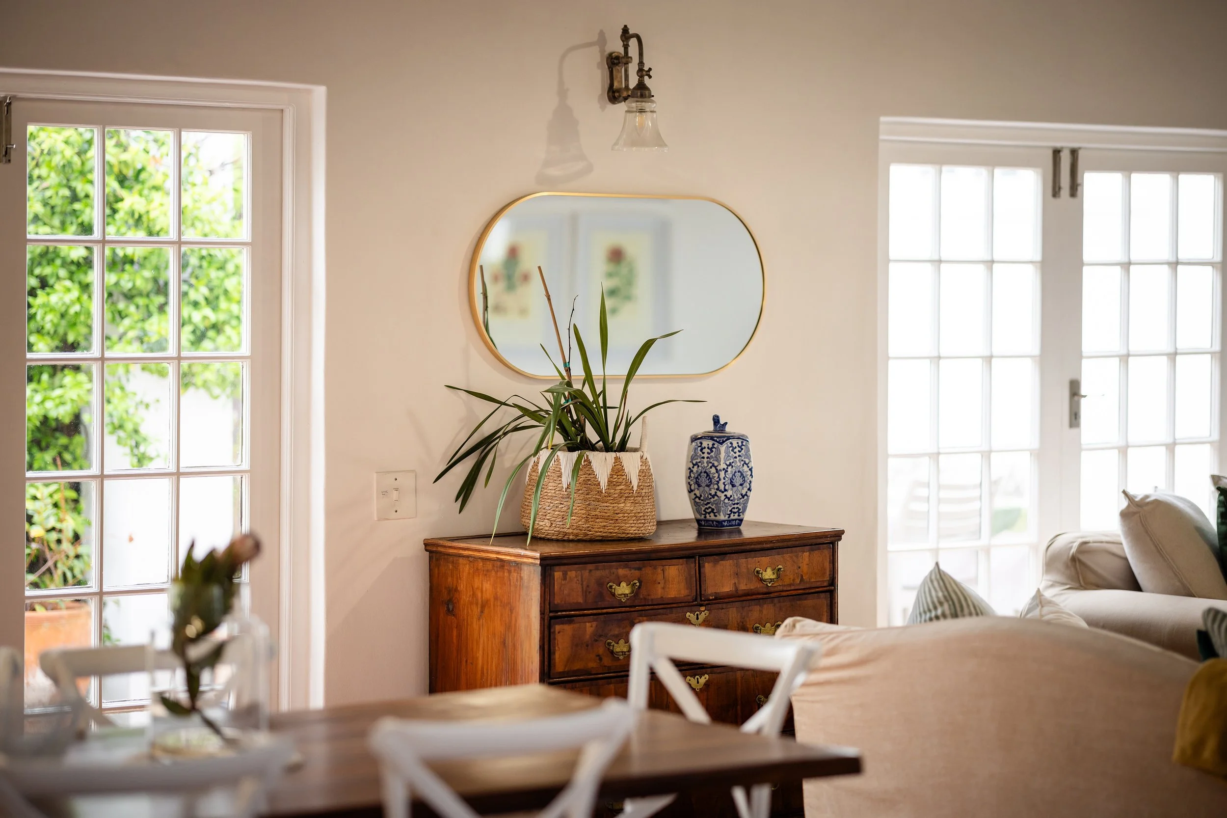 A cozy living room corner with a wooden sideboard, a potted plant, a mirror, and large windows allowing natural light.
