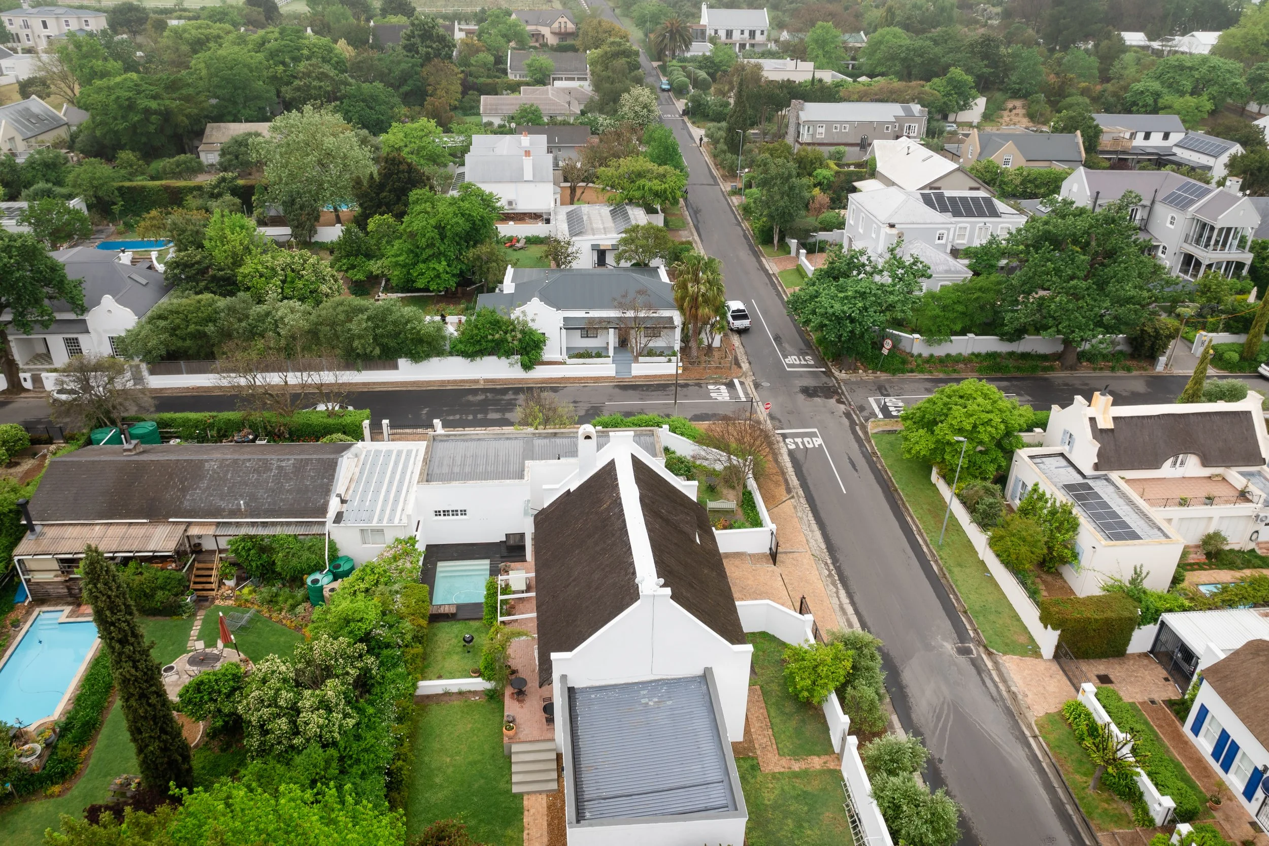 An aerial view of a suburban neighborhood with houses, trees, streets, and a swimming pool in the backyard. The neighborhood is lush with greenery and organized with various homes featuring different architectural styles.