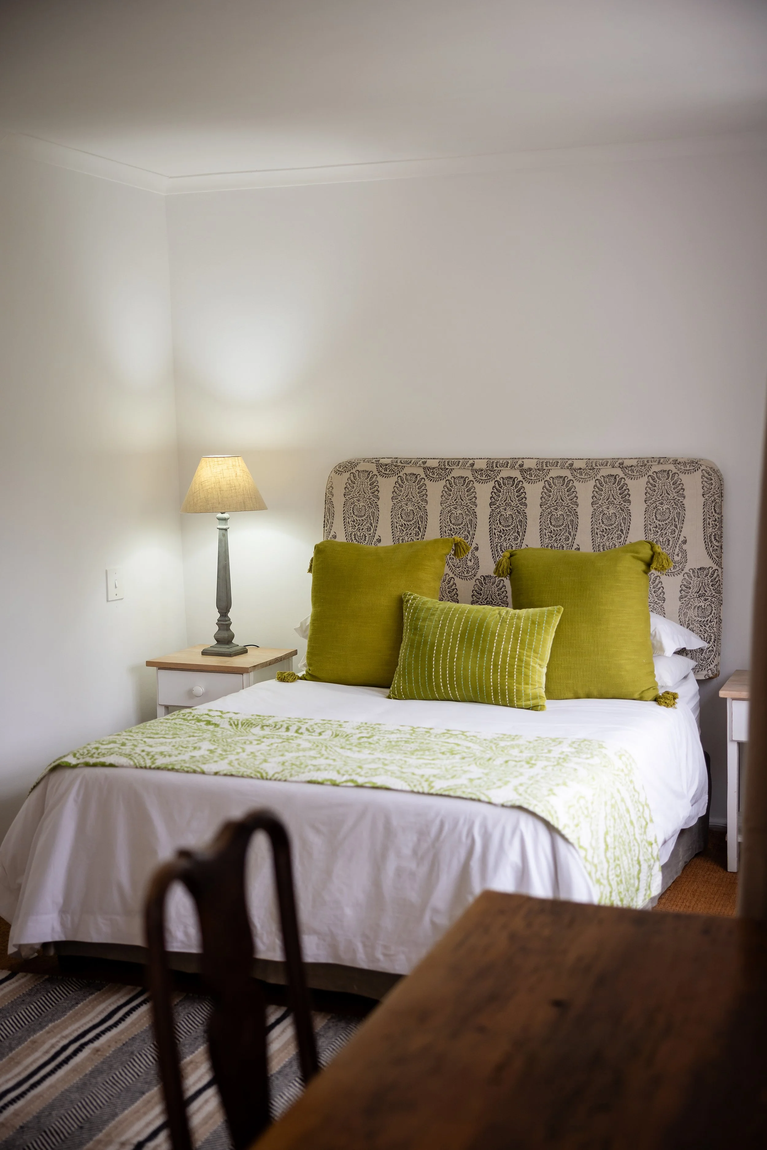 A bedroom with a bed that has a patterned headboard, green pillows, and a white and green bedspread. There is a nightstand with a lamp on the left side and part of a wooden chair or table in the foreground. The wall behind the bed is plain and light-colored.