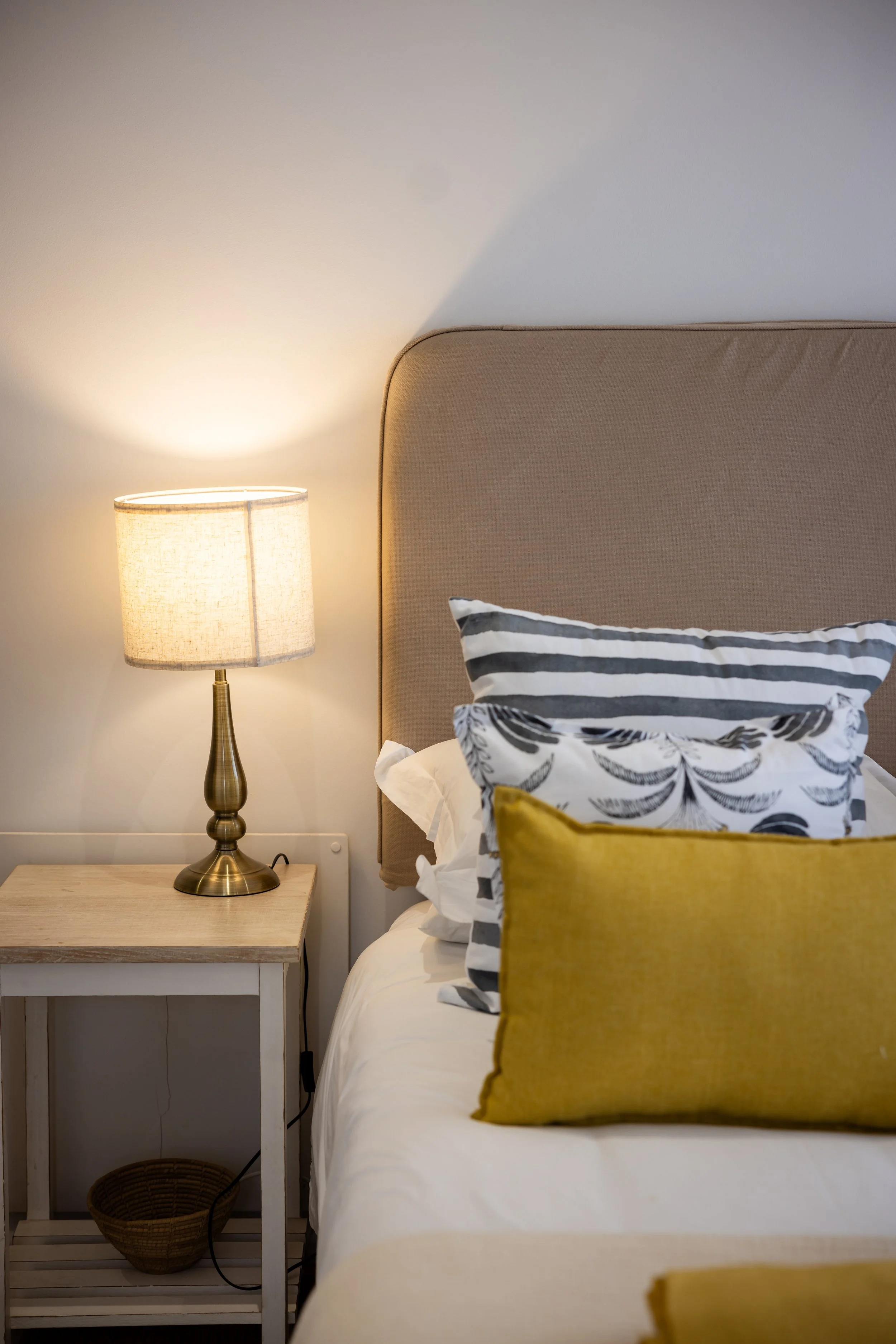 A bedroom corner showing a beige upholstered headboard, a bedside table with a brass lamp, and pillows with black, white, and yellow pillowcases.