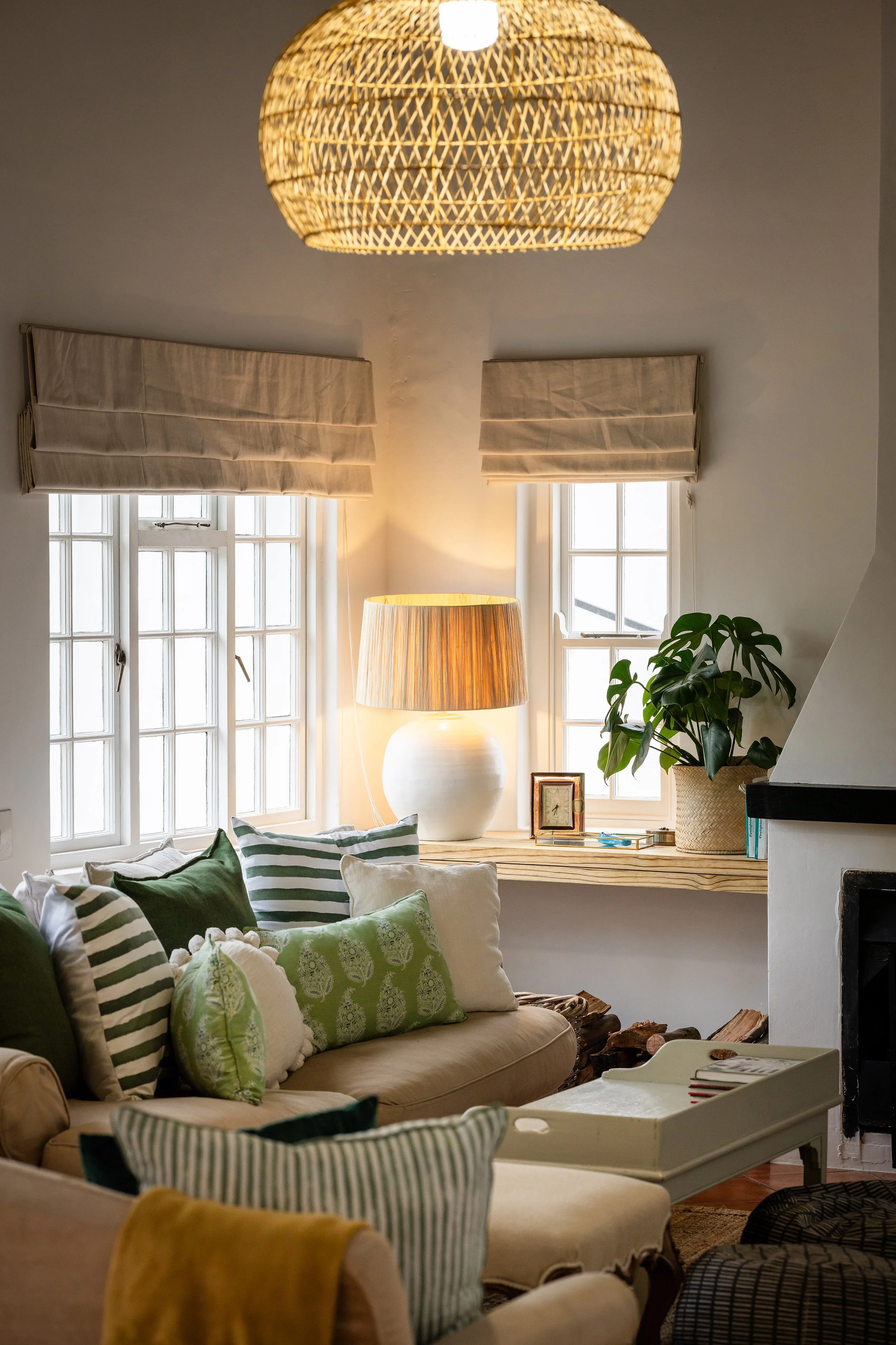 Cozy living room with beige sofas, assorted green and striped pillows, large windows with beige Roman shades, a wicker ceiling light, a large white ceramic lamp, a houseplant in a woven basket, and wooden logs on the floor.