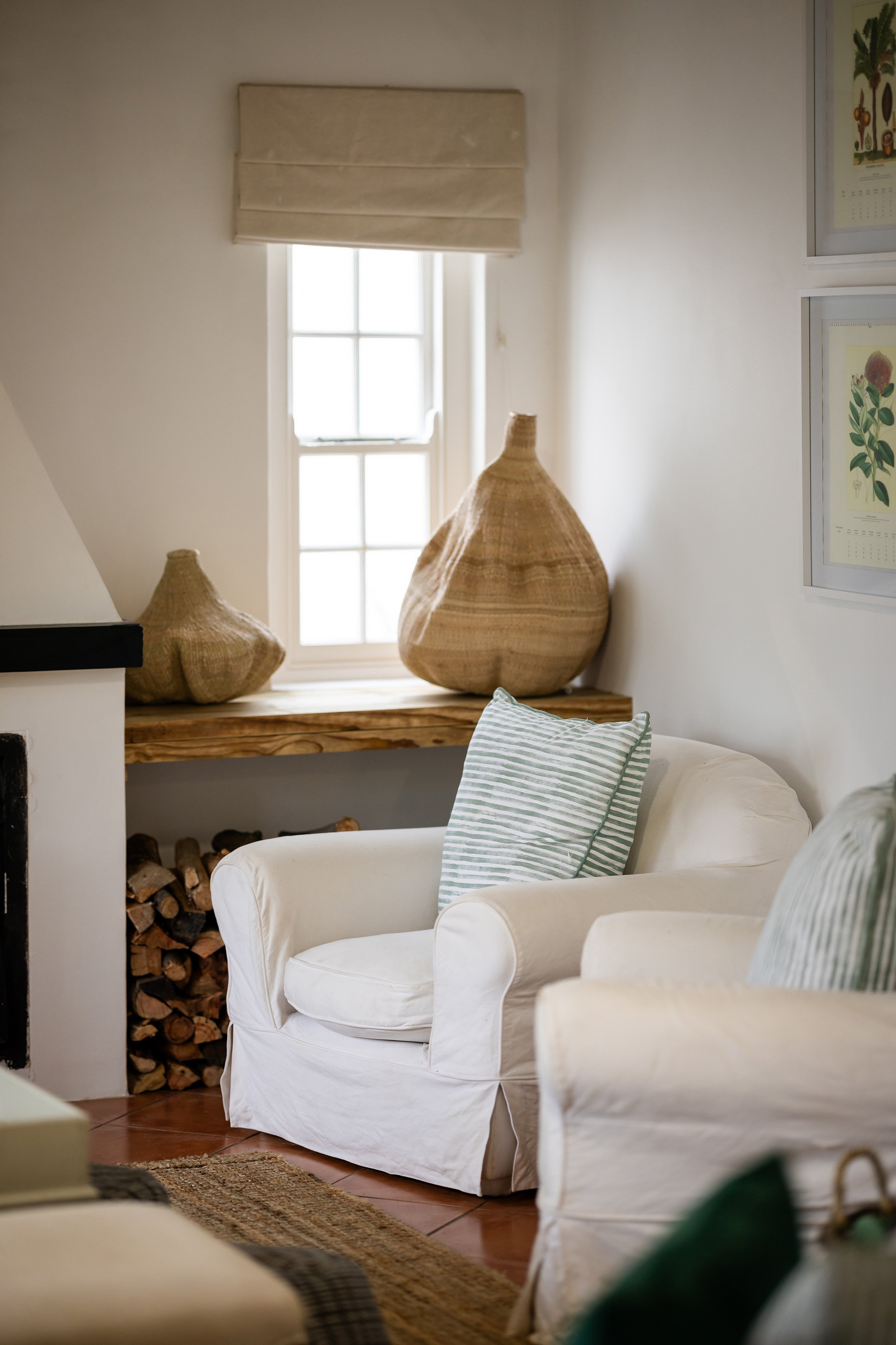 Living room with white armchairs, striped pillows, a window with a beige Roman shade, woven vases on a wooden shelf, and framed botanical prints on the wall.