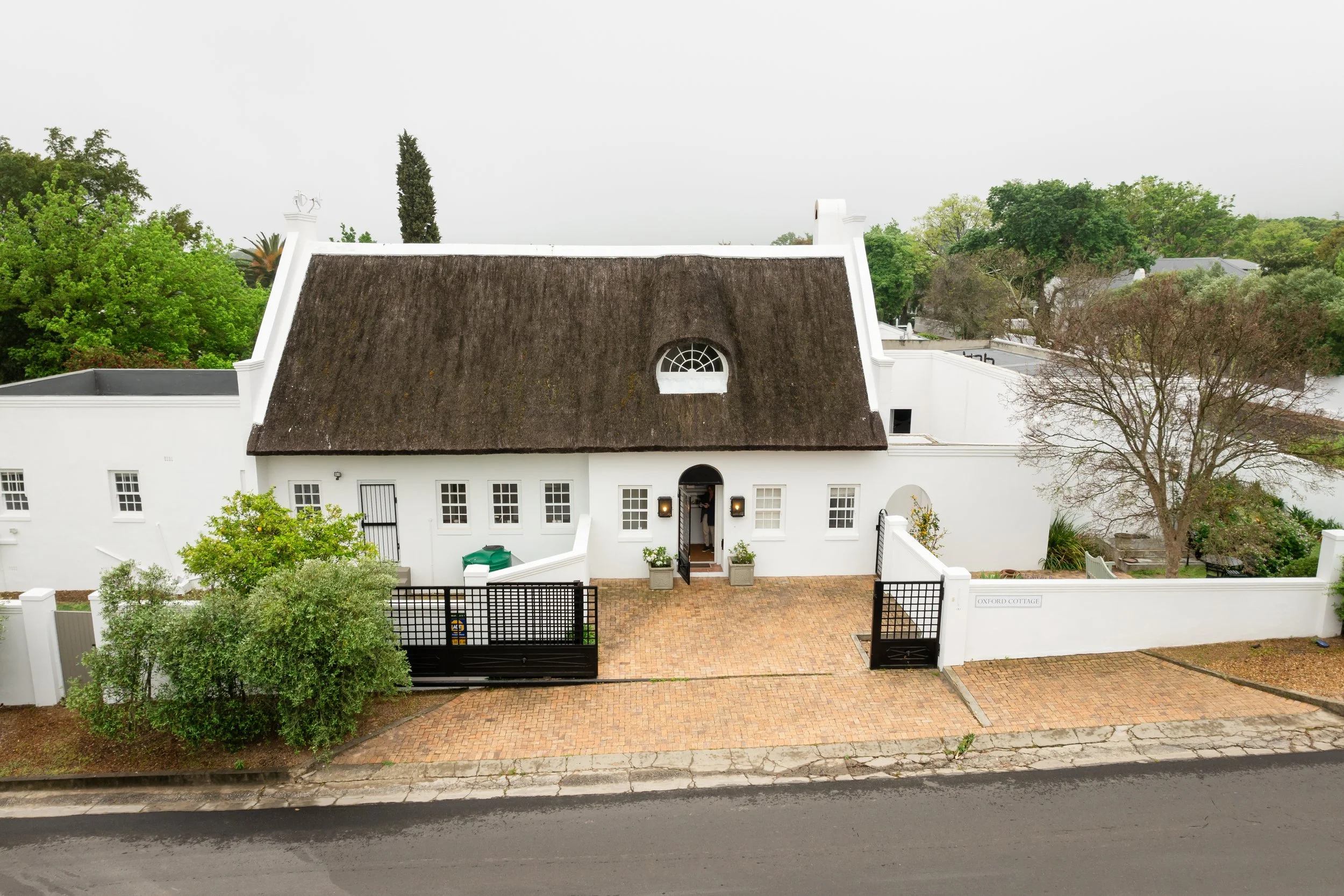 Front view of a white cottage with a thatched roof, a small arched window, and a person standing in the doorway, surrounded by trees and a brick-paved yard, with a black gate and fence.