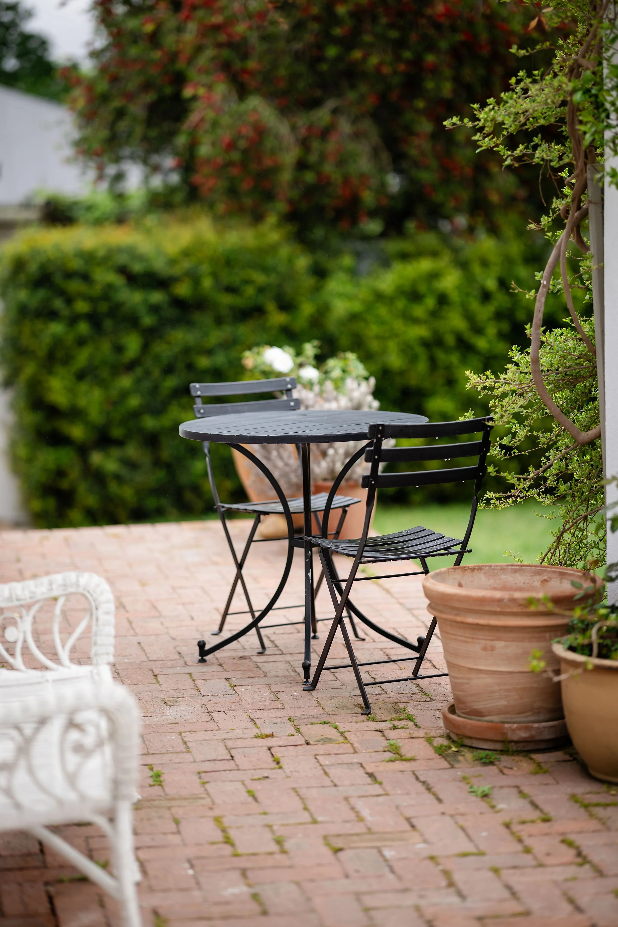 A small round black metal table with two matching chairs on a brick patio, surrounded by large terracotta pots with plants, and a lush green hedge in the background.