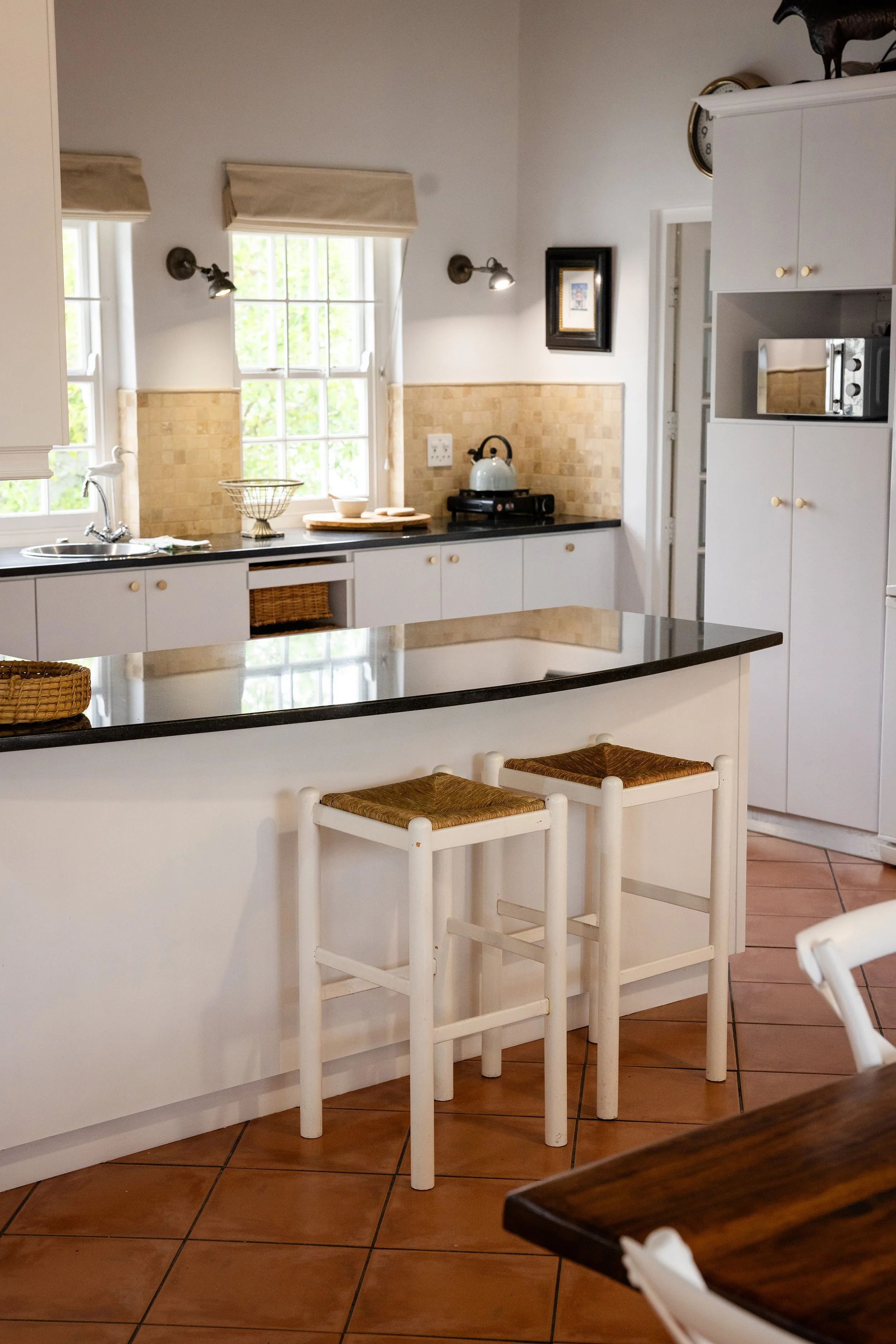 A kitchen scene with white cabinetry, beige tile backsplash, and a black countertop. There are two white chairs with woven seats in the foreground. A window with blinds and two wall-mounted lights are visible, along with various kitchen items including a kettle, a basket, and a microwave.