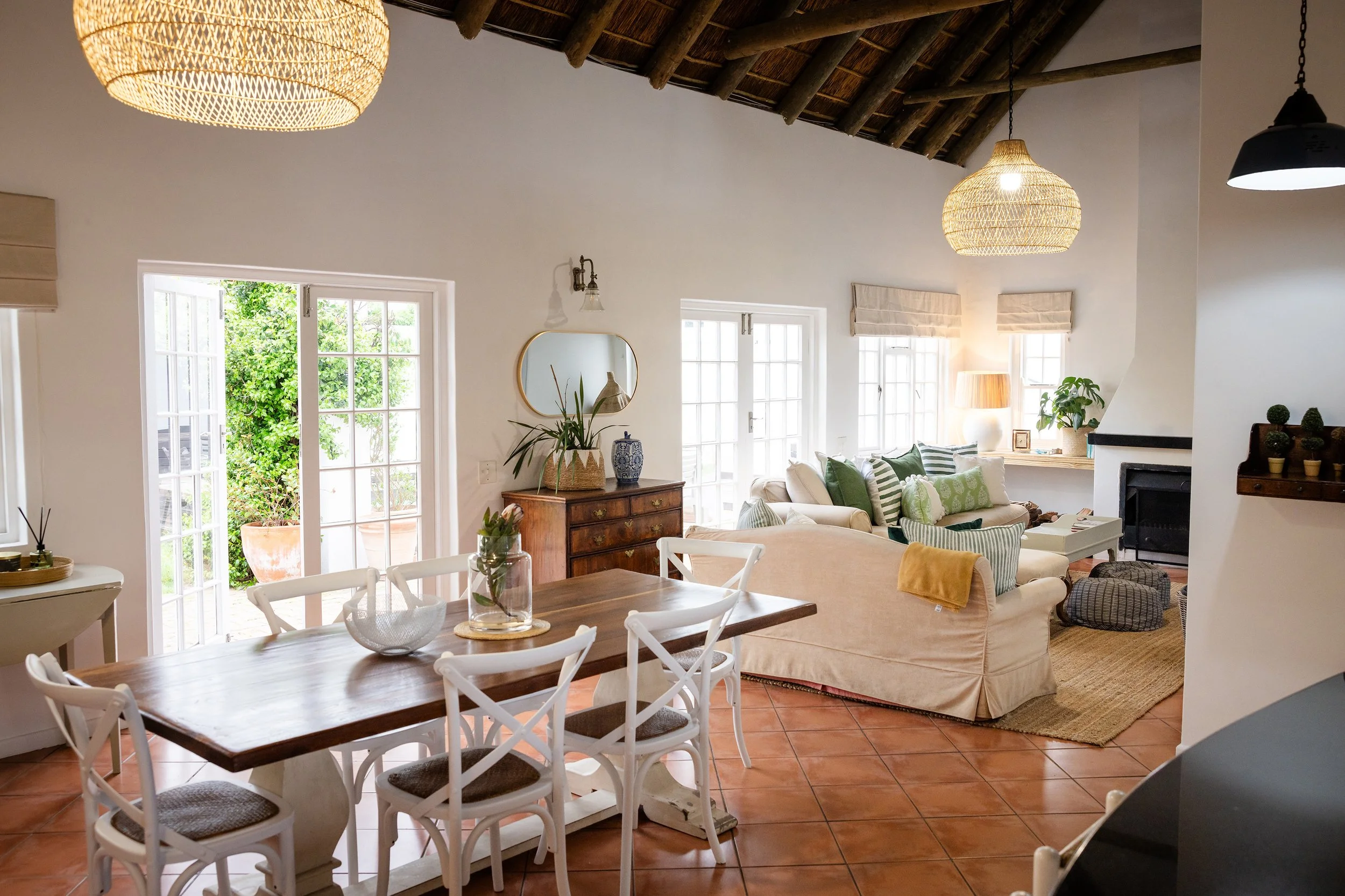 A cozy living room with white walls, wooden furniture, a dining table with white chairs, and a sitting area with a beige sofa. Large windows and glass doors bring in natural light, with greenery visible outside. There are hanging woven pendant lights and various decorative plants inside.