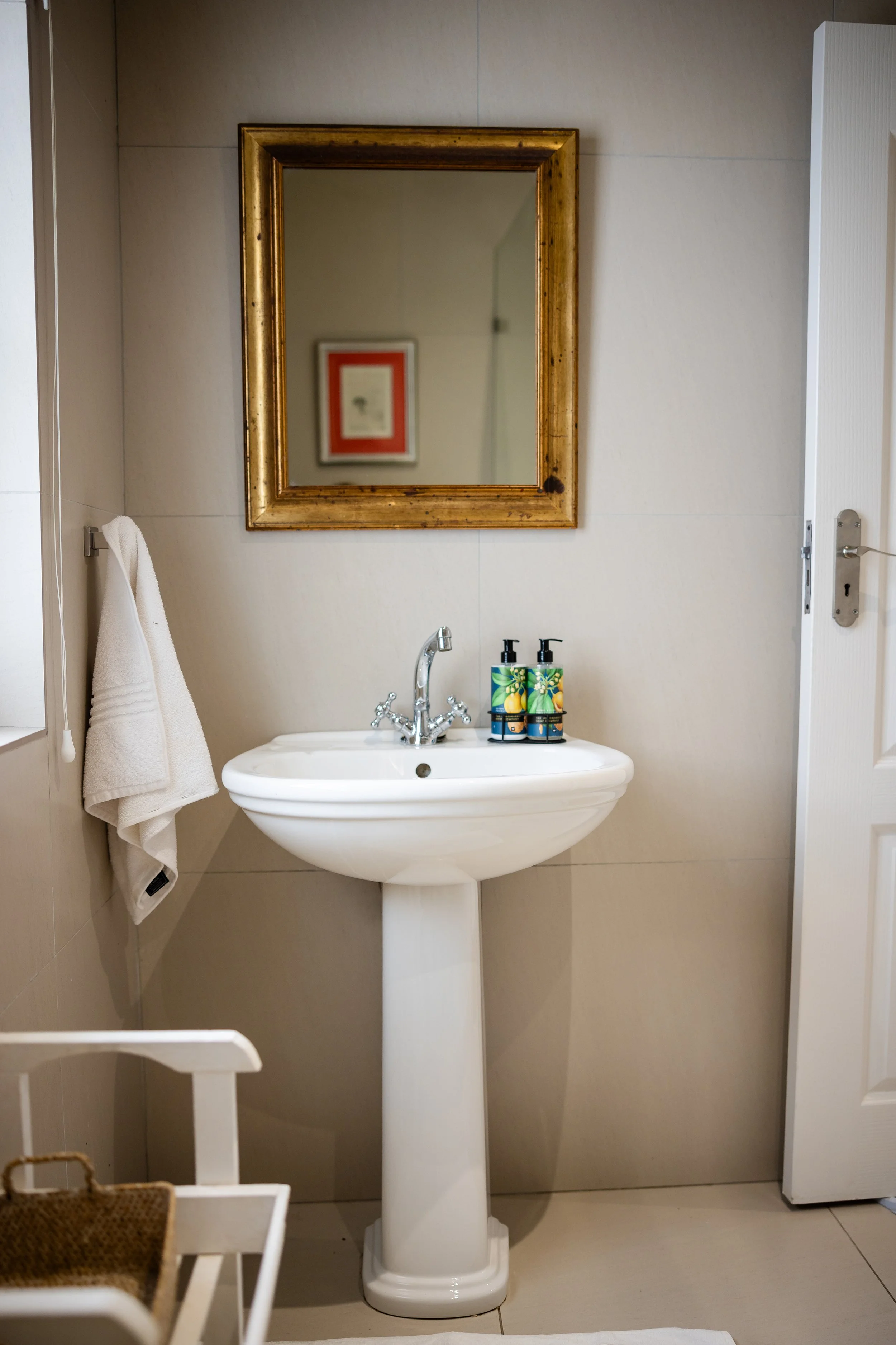 A bathroom with a white sink on a pedestal, a gold framed mirror above it, and two soap dispensers on the sink. To the left, a white towel hangs on a hook, and there is a white chair in front of the sink. Part of the door with a latch is visible on the right.