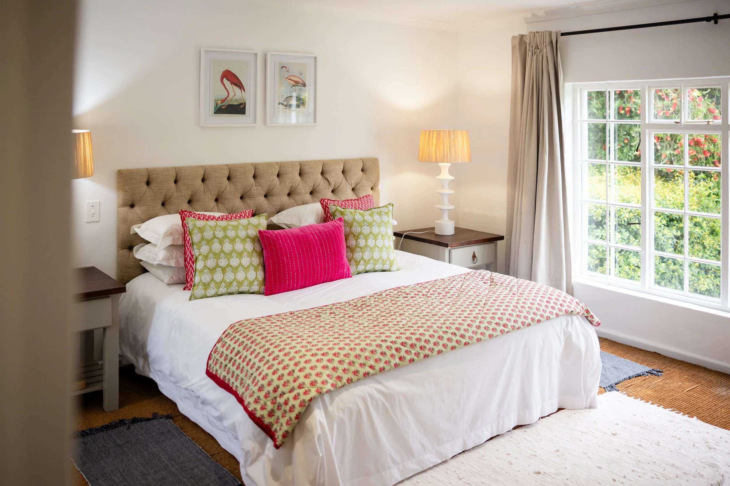 A cozy bedroom featuring a bed with a beige tufted headboard, colorful pillows, and a patterned quilt. There are two white nightstands with lamps, a large window with curtains, and framed bird artwork on the wall.