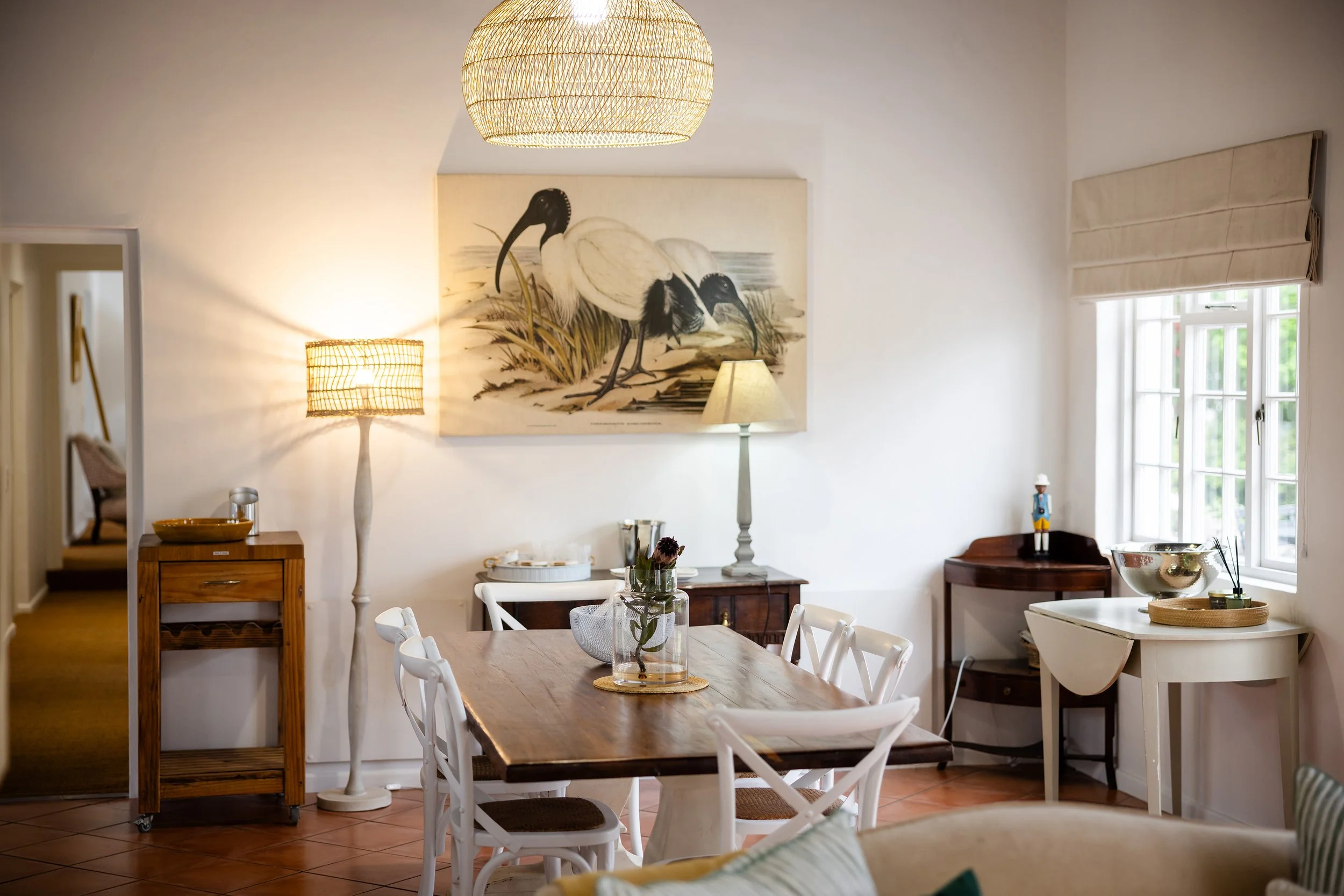 A cozy dining room with a wooden table surrounded by white chairs, a large painting of two white herons hanging on the wall, two standing lamps, a window with beige Roman shades, and various decorative items like a glass vase with flowers and a bowl.