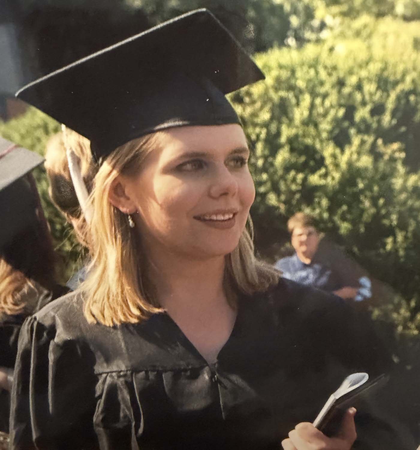 Sonia Huntsman in a graduation cap and gown, smiling outdoors during sunset, with trees in the background, holding a booklet or program.
