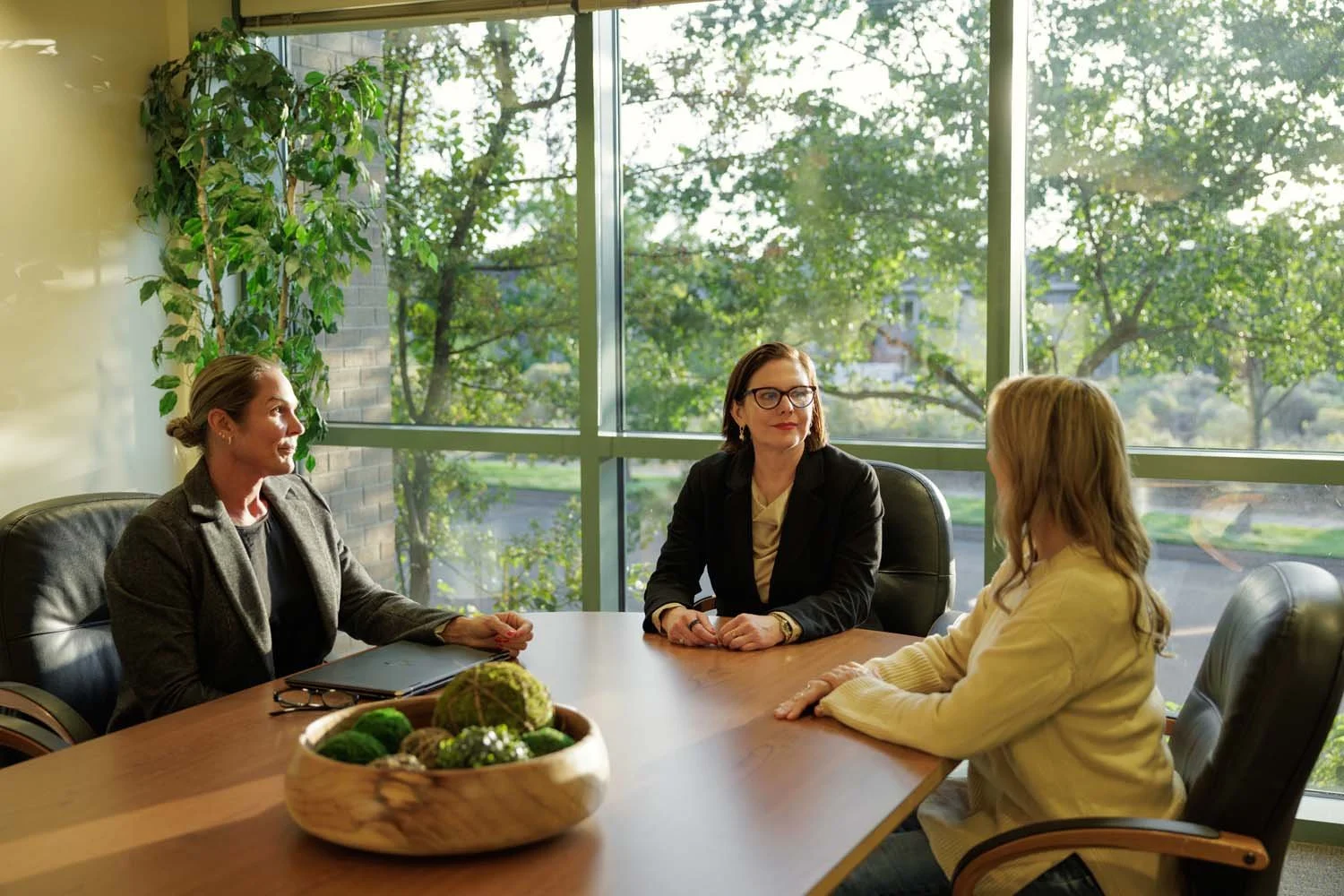 Sonia Huntsman and two women having a discussion in a corporate office conference room with large windows showing trees outside. There is a bowl of decorative green and brown balls on the table.