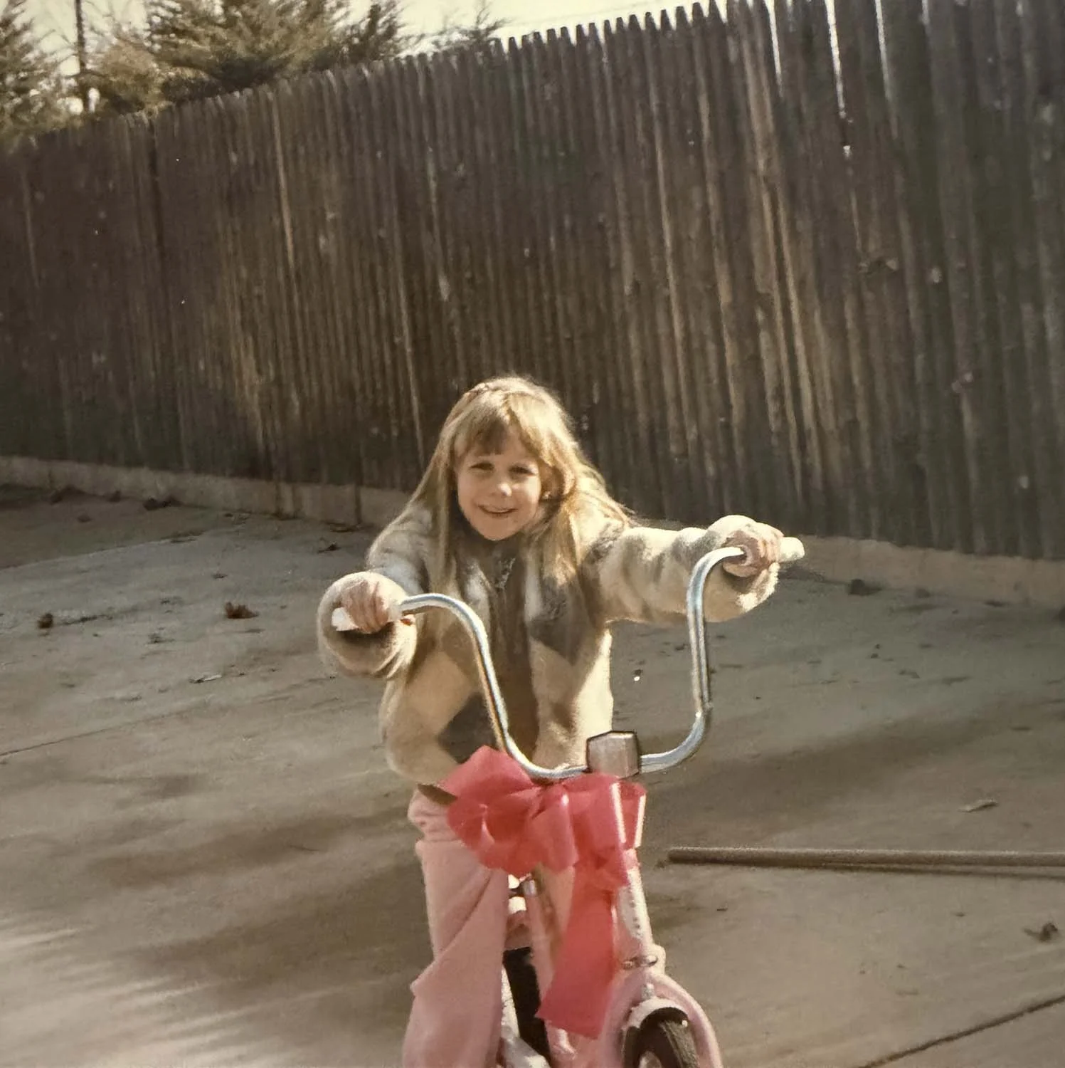 Young Sonia with long light brown hair smiling and riding a pink bicycle with a big pink bow. She is wearing a beige coat and pink pants, outdoors at night on a street with a tall wooden fence in the background.